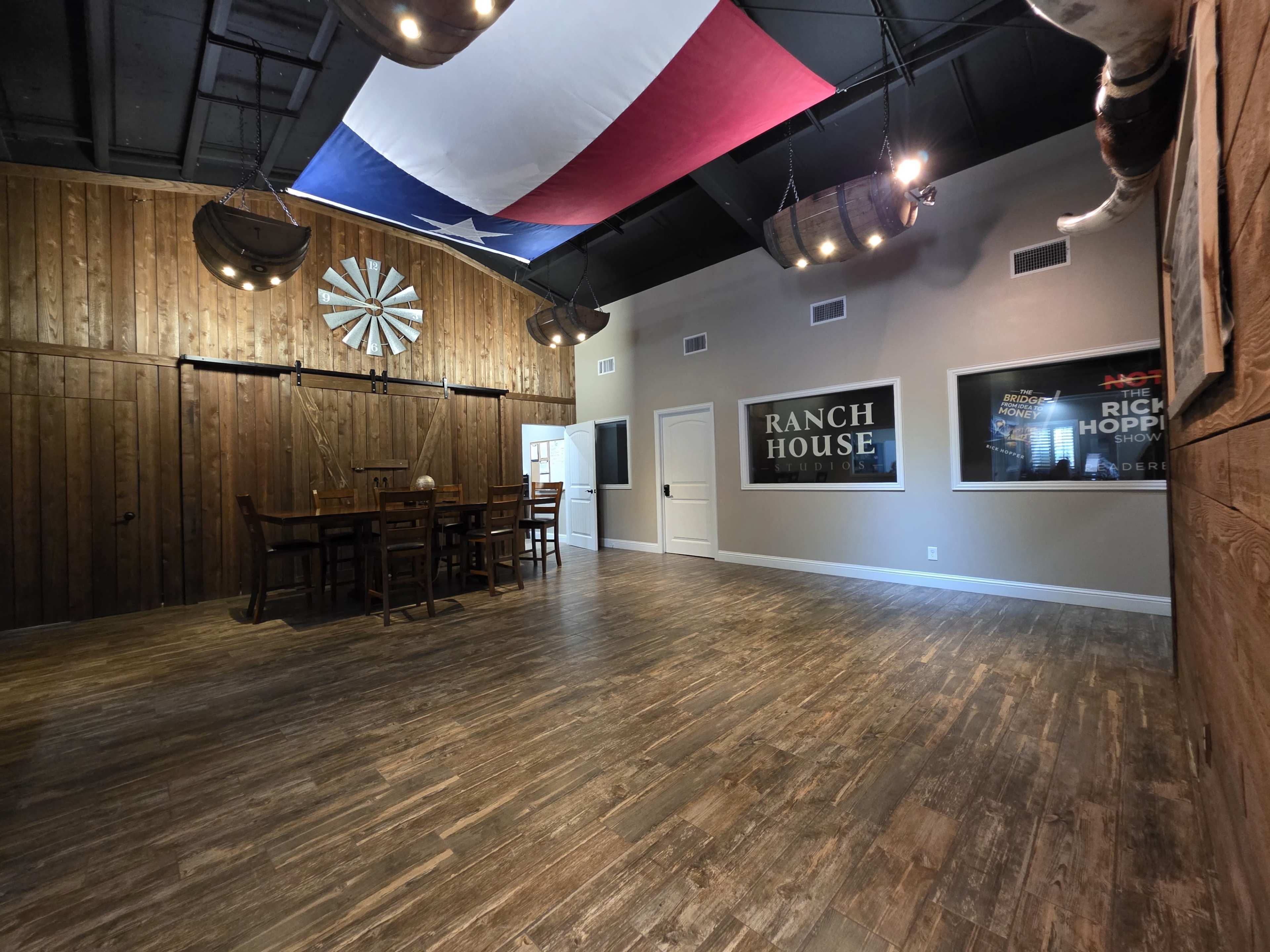 The image shows an interior space with wooden walls, a Texas flag hanging from the ceiling, a large dining table with chairs, and a few framed posters on the wall.