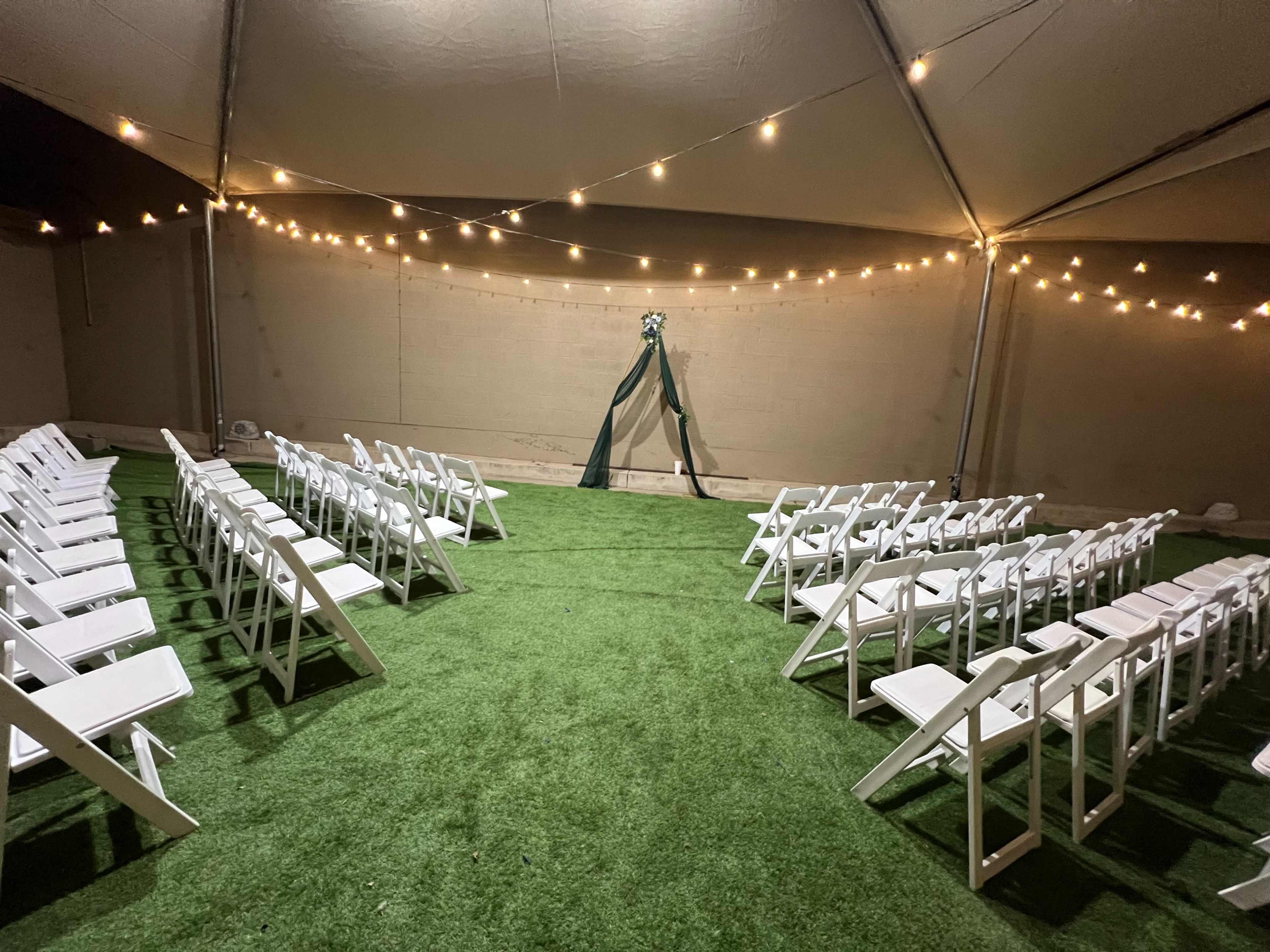 A tented wedding setup features neatly arranged white chairs on artificial grass facing a decorated arch under string lights.
