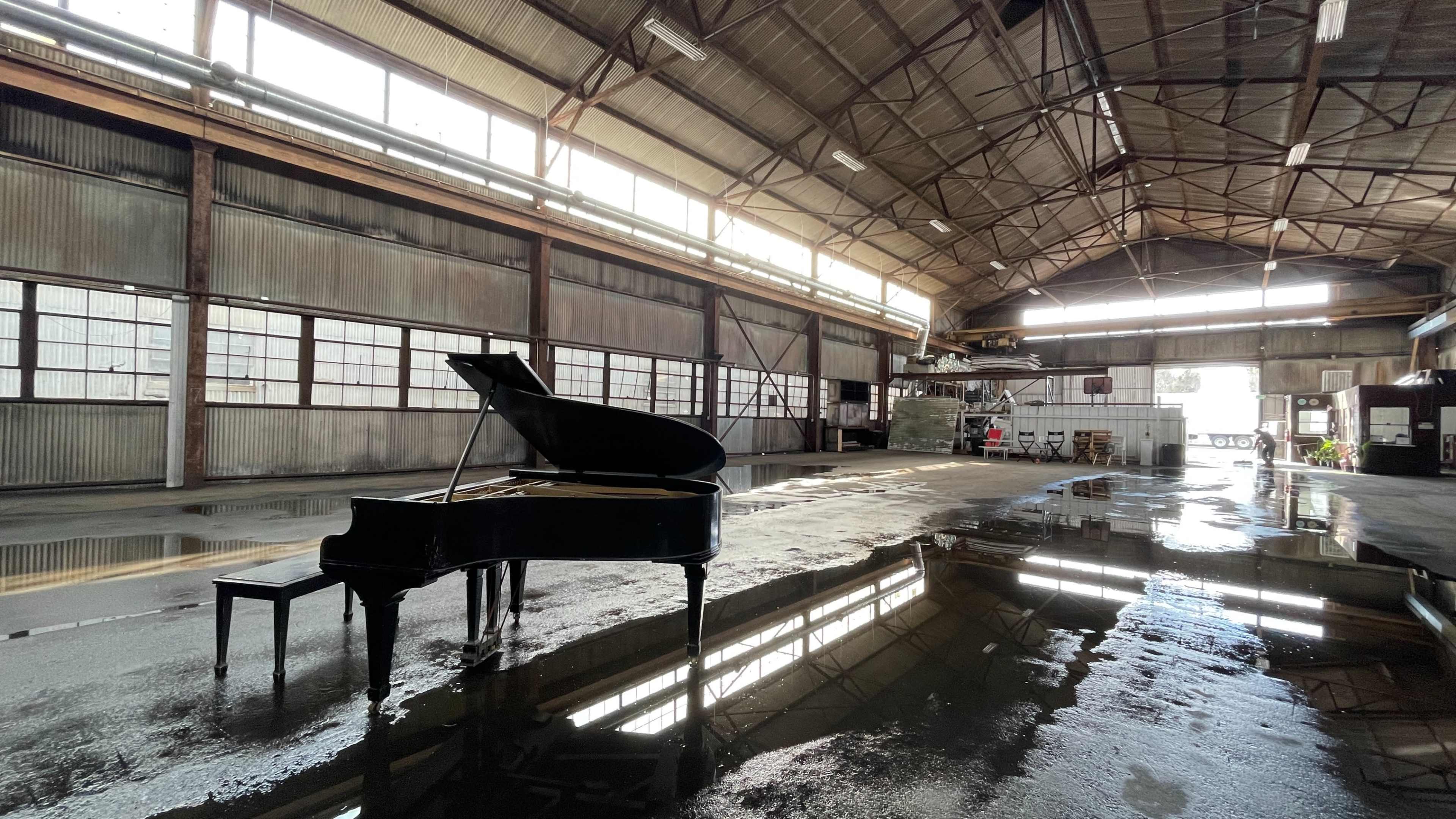 A grand piano sits in the center of a large, industrial space with water pooled on the floor reflecting its image.