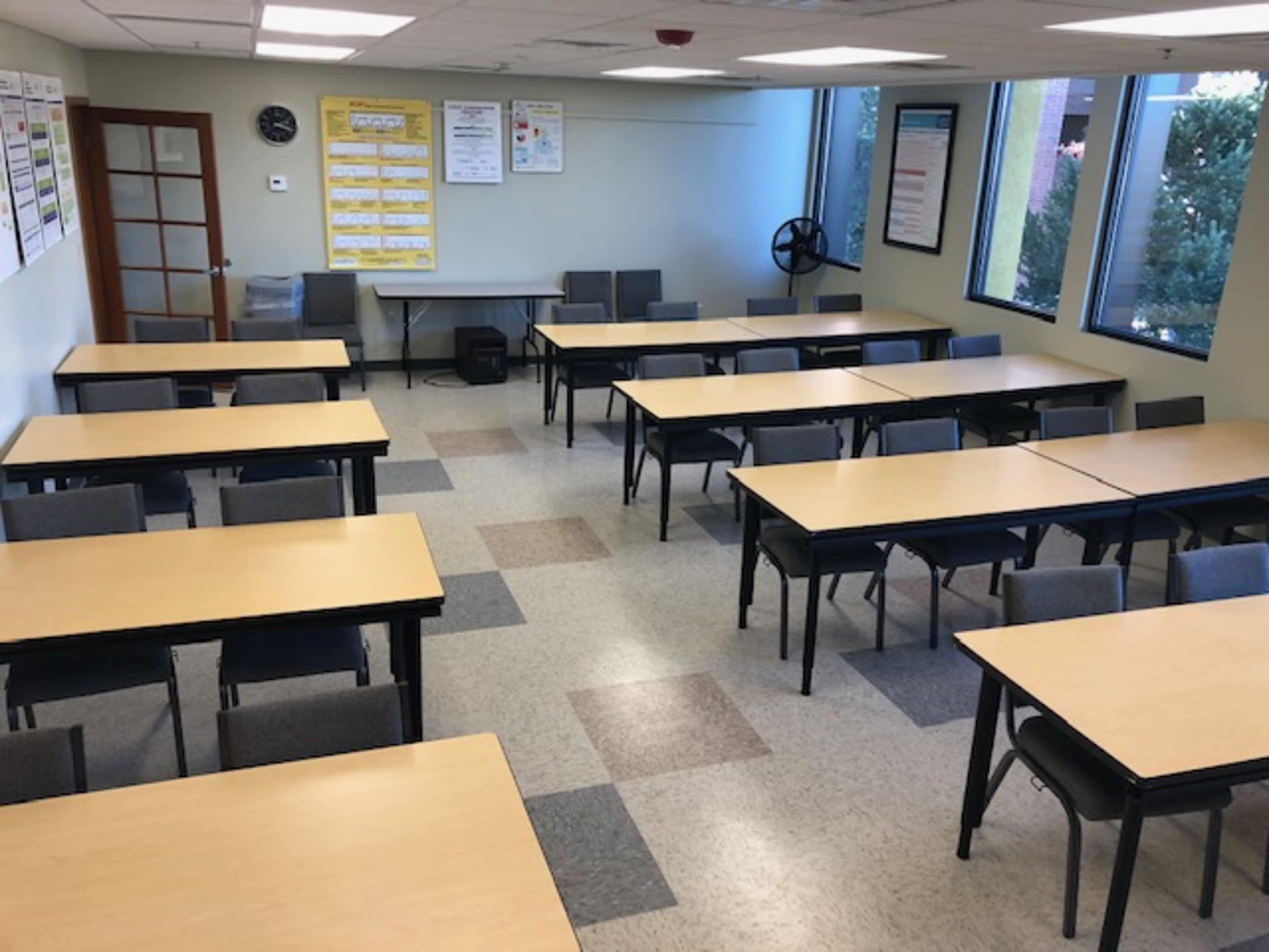 A classroom is arranged with multiple tables and chairs, with posters on the walls and large windows letting in natural light.