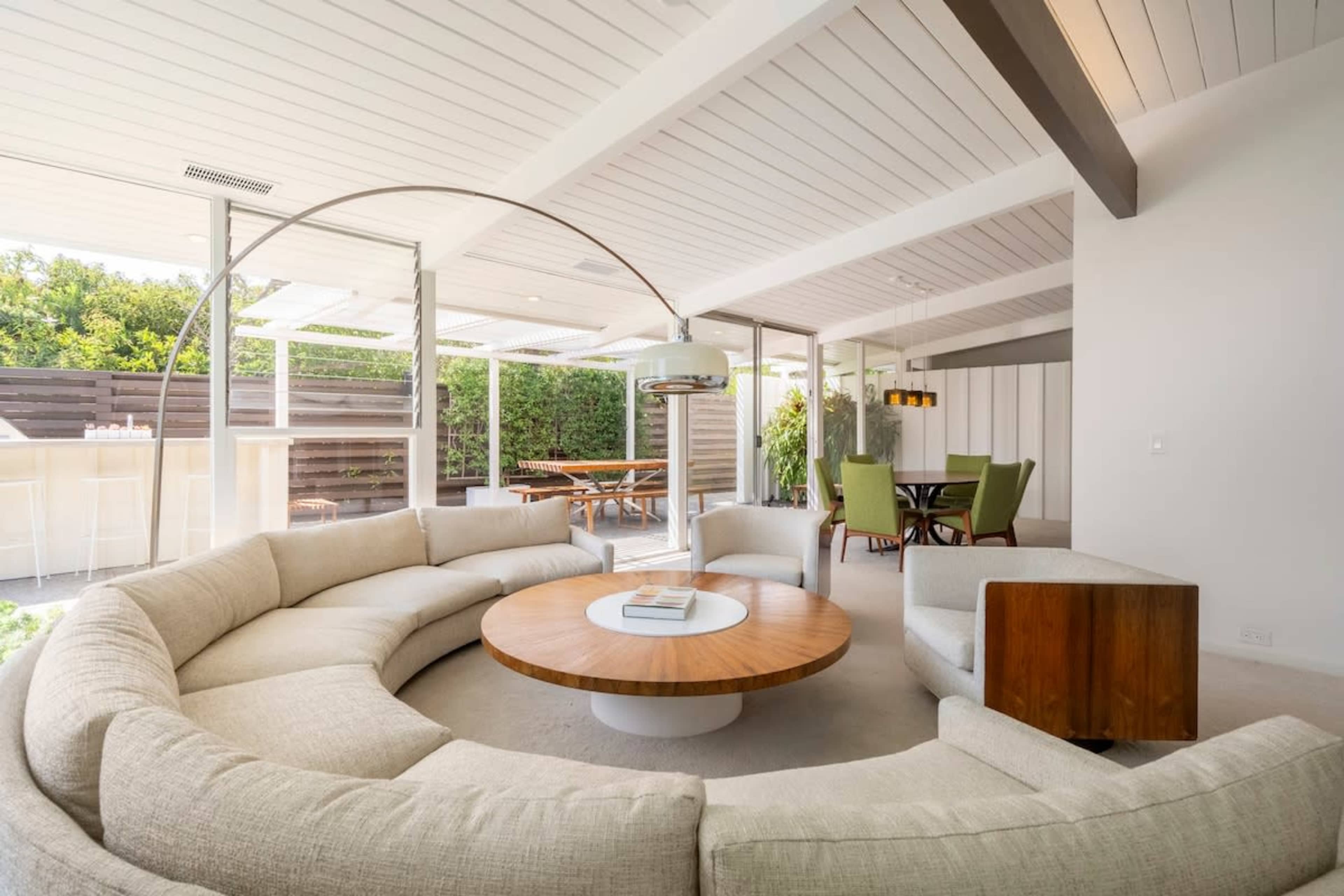 The image shows a modern, open-plan living space featuring a circular sofa with a wooden table in the center, surrounded by large windows and views of an outdoor patio.