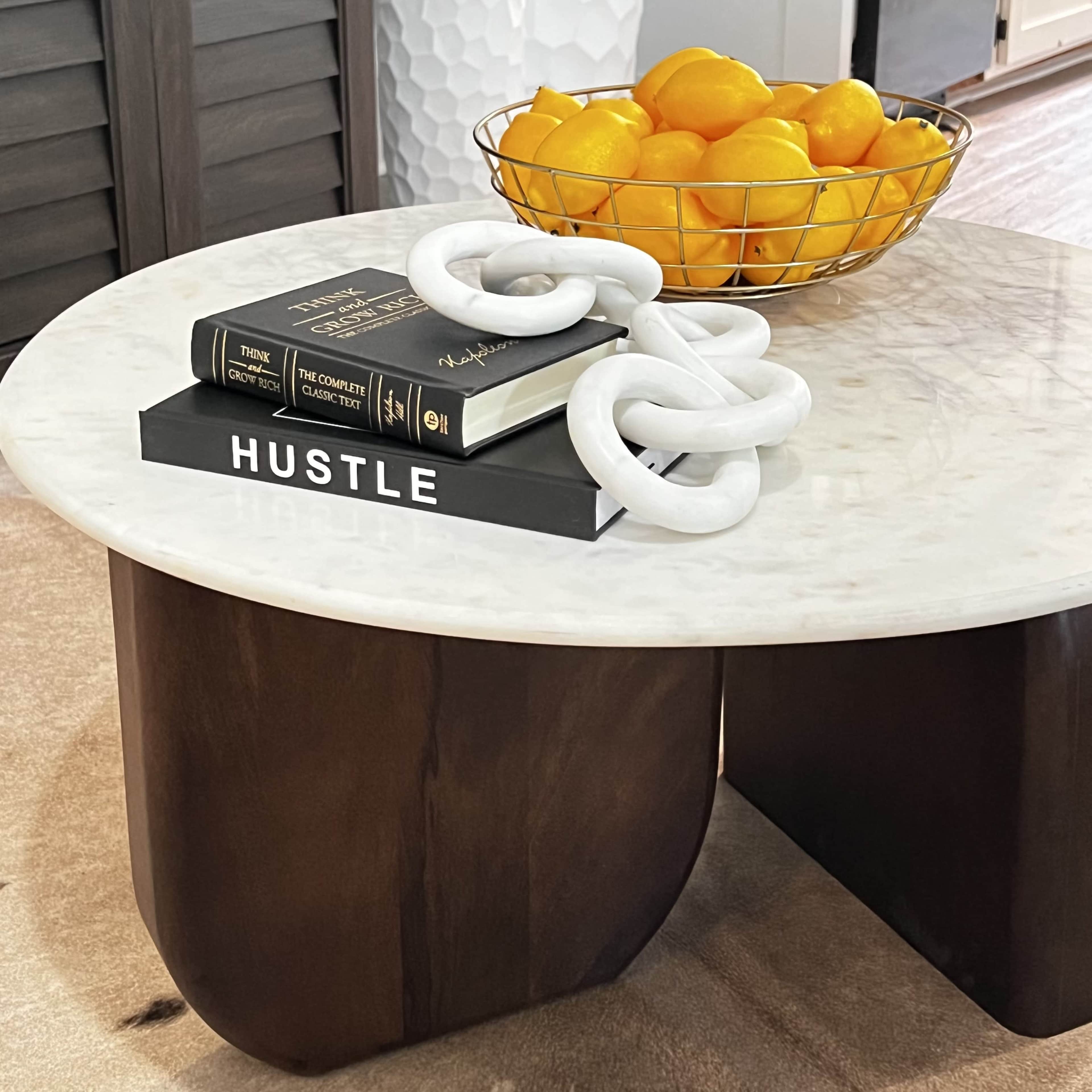 A round coffee table made of dark wood holds a stack of books titled "HUSTLE" and "The Complete Collections," along with a decorative chain and a bowl of lemons.