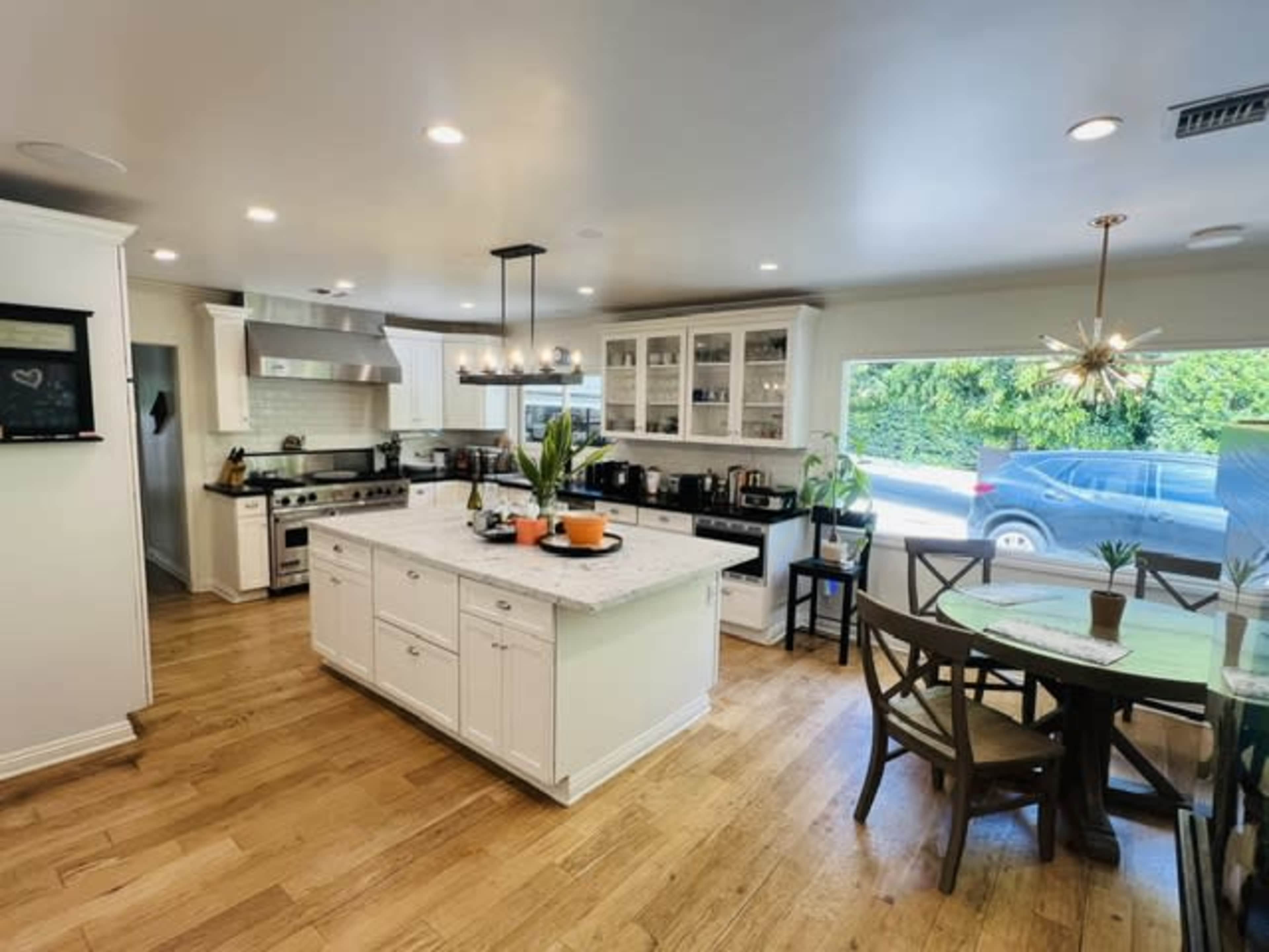 The image shows a modern kitchen with a central island, wooden floors, and a large window providing natural light.
