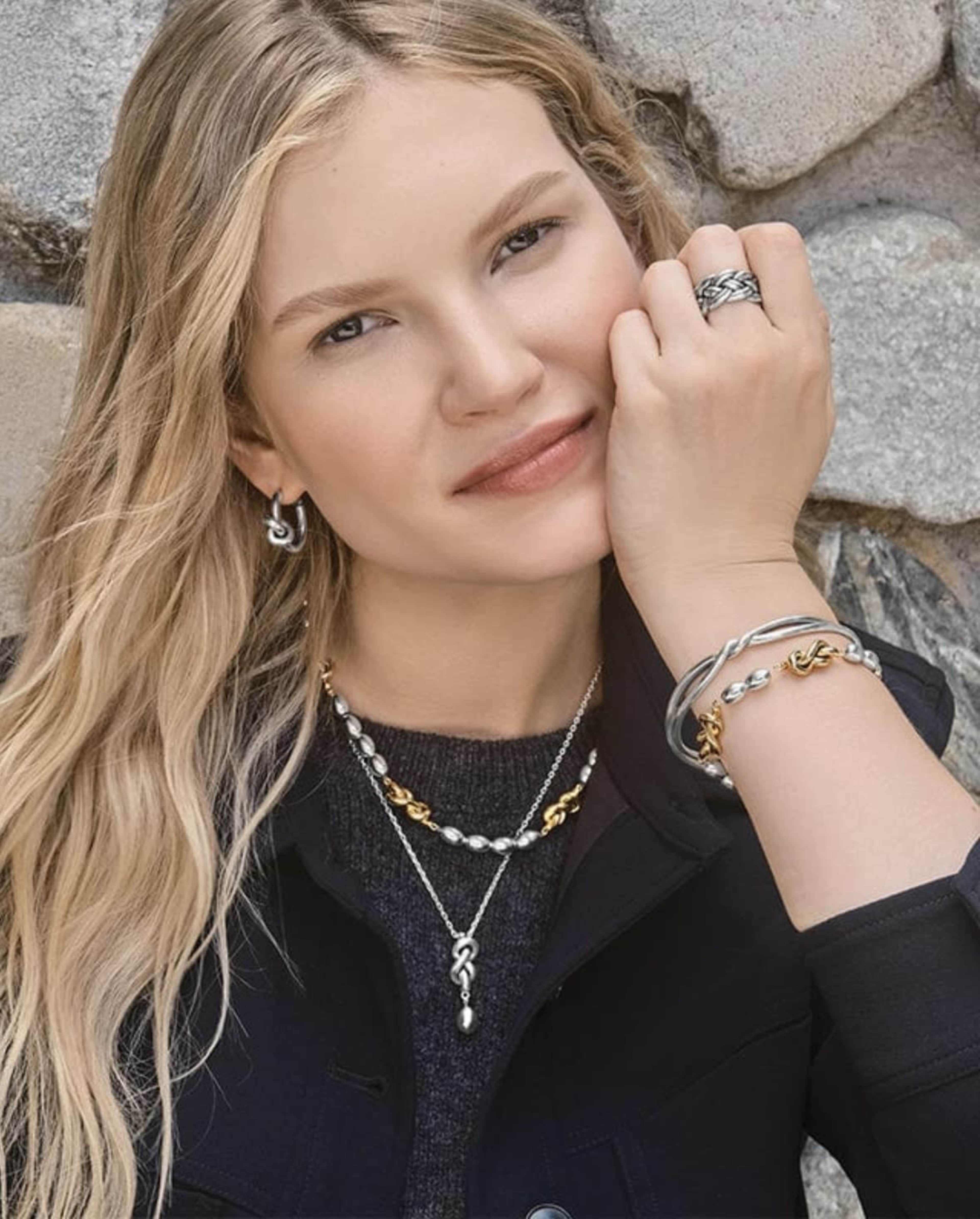 A woman poses against a stone wall while showcasing various pieces of jewelry on her neck and wrists.