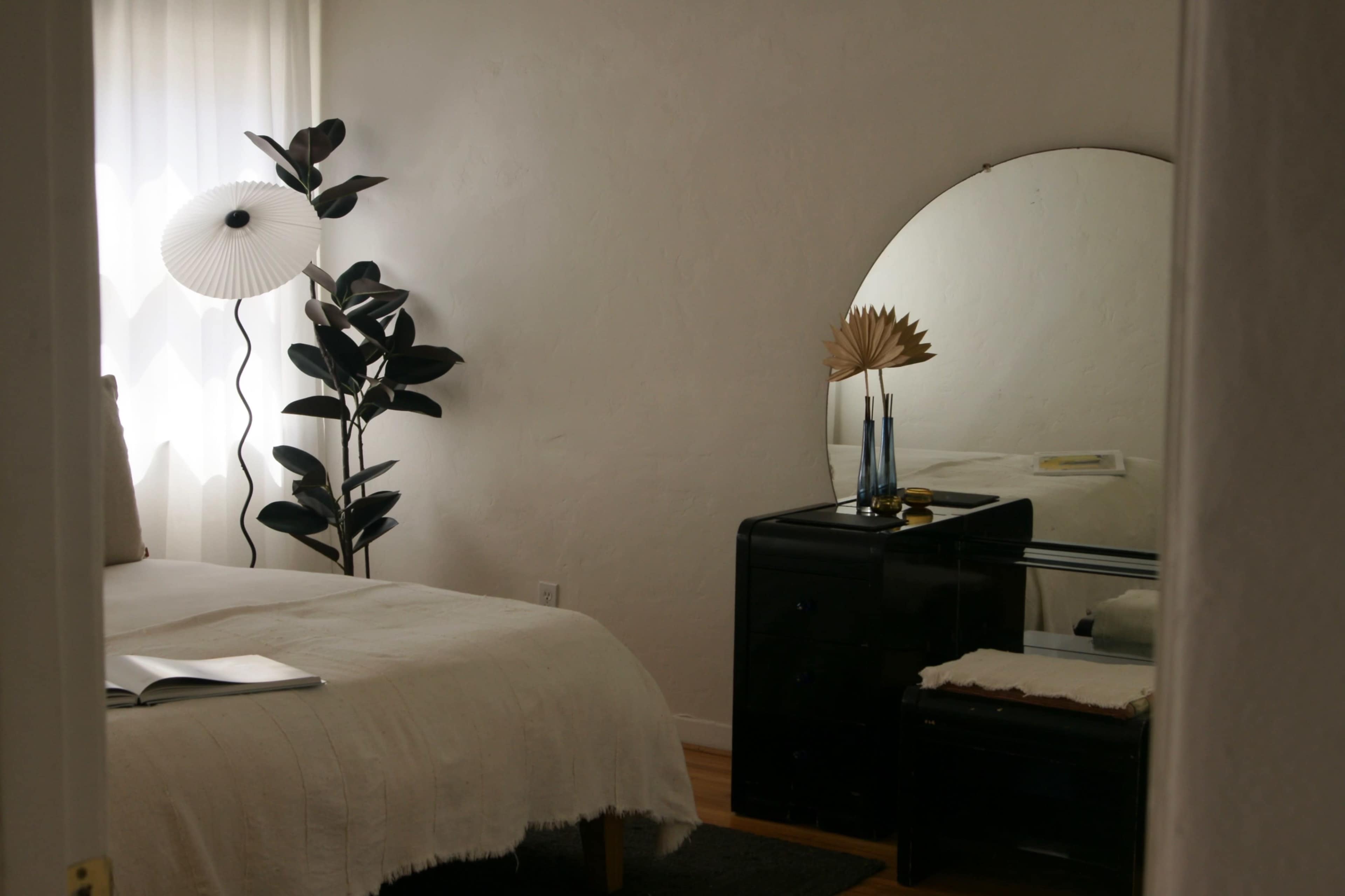 The image shows a simple bedroom featuring a bed with a white cover, a mirror, a black dresser, and a potted plant.