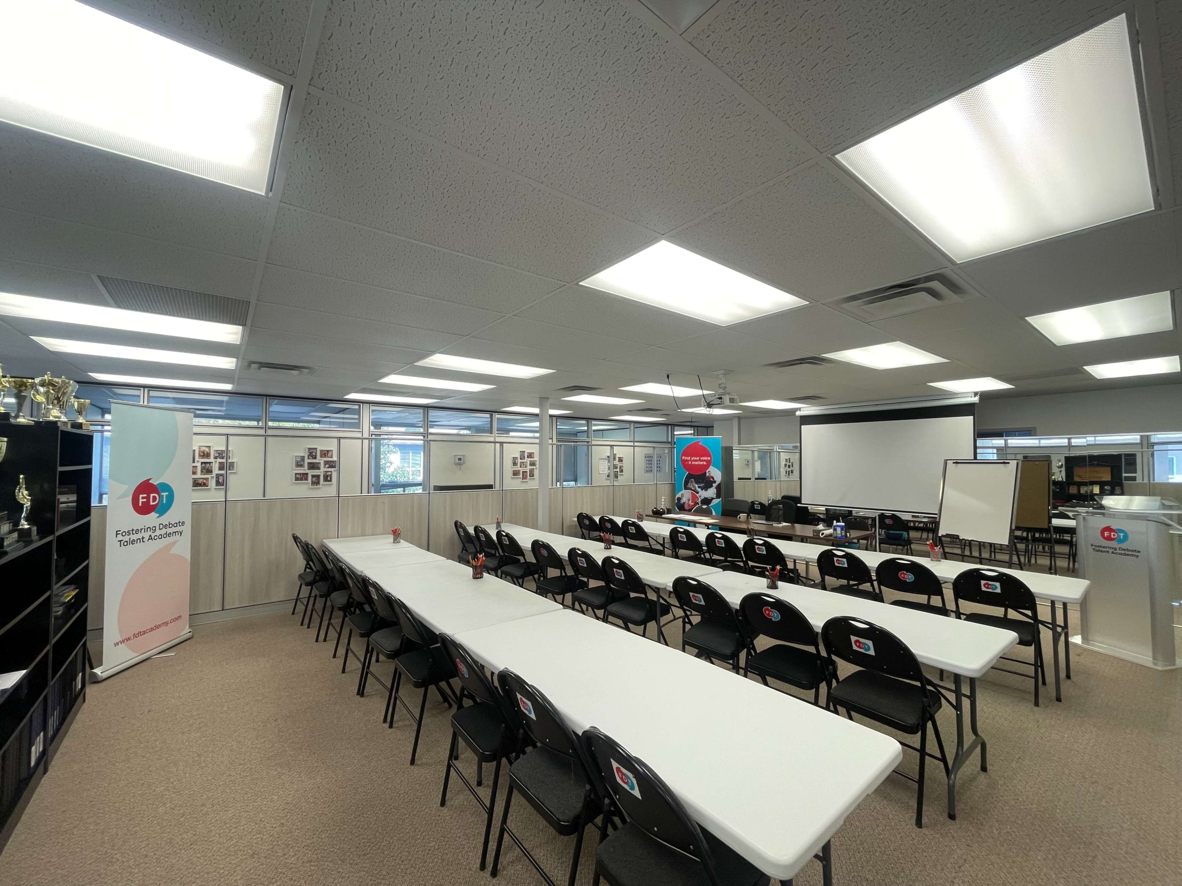 A clean conference room arranged with rows of white tables and black chairs, equipped with a projector screen and presentation tools.