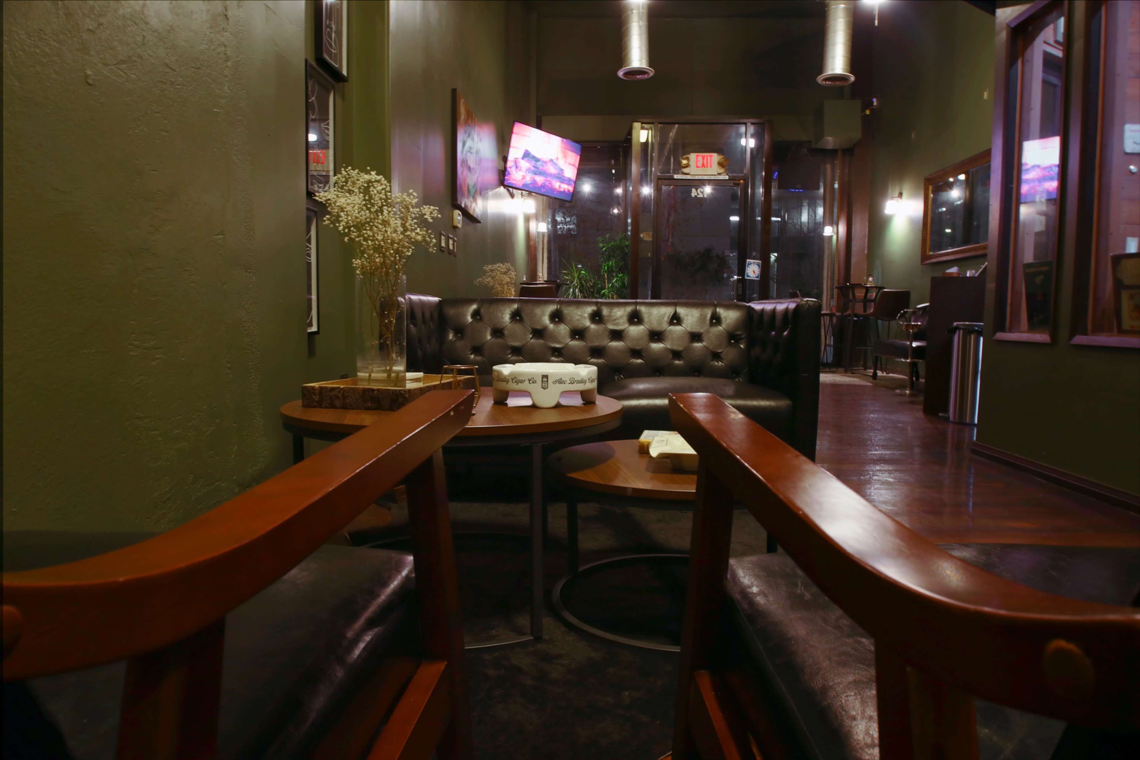 A cozy lounge area features a dark leather sofa, wooden chairs, and a side table in a green-walled space with warm lighting.