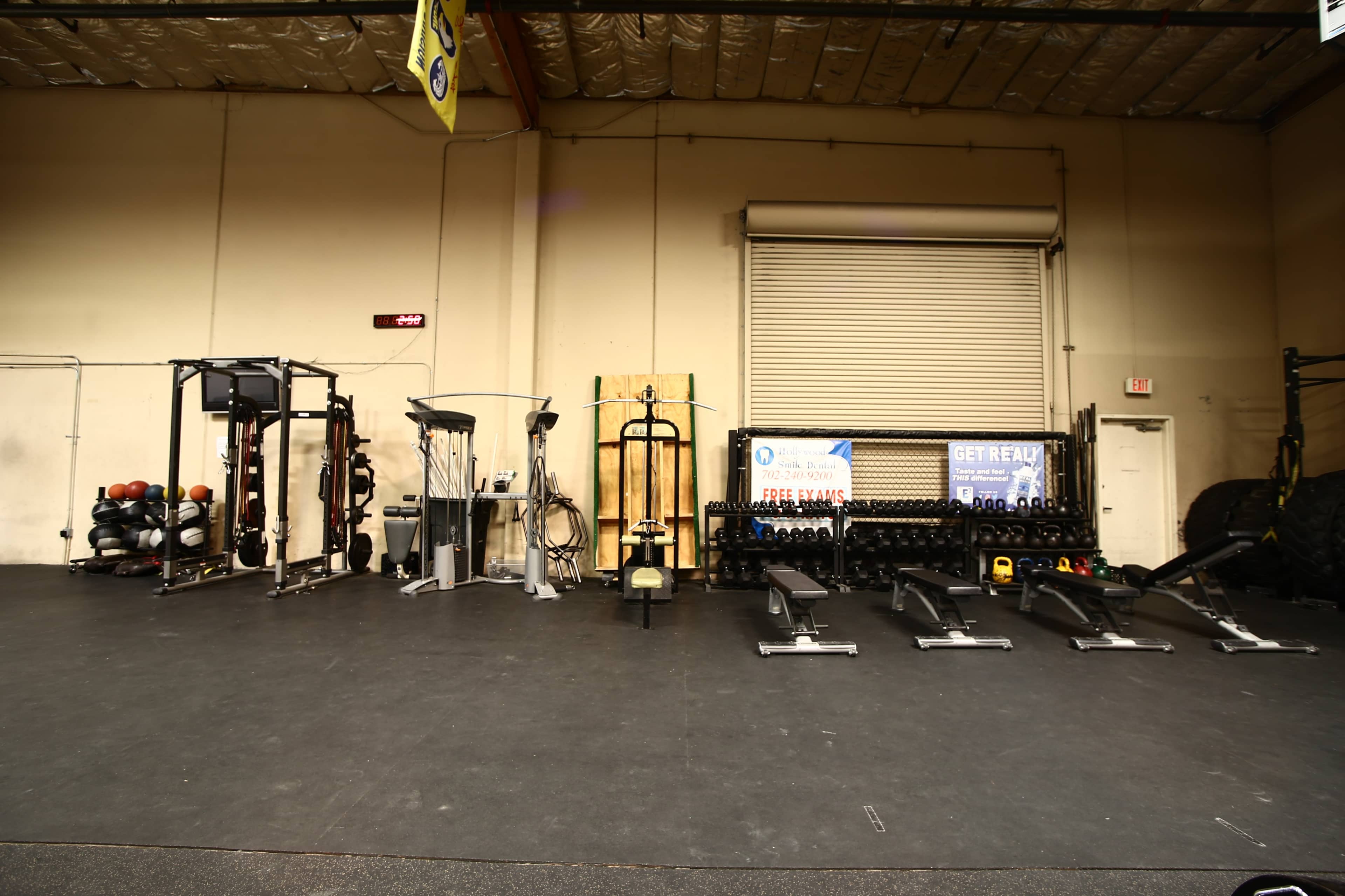 A gym interior with various exercise equipment, including weight racks, rowing machines, and strength training apparatus, arranged along the walls.