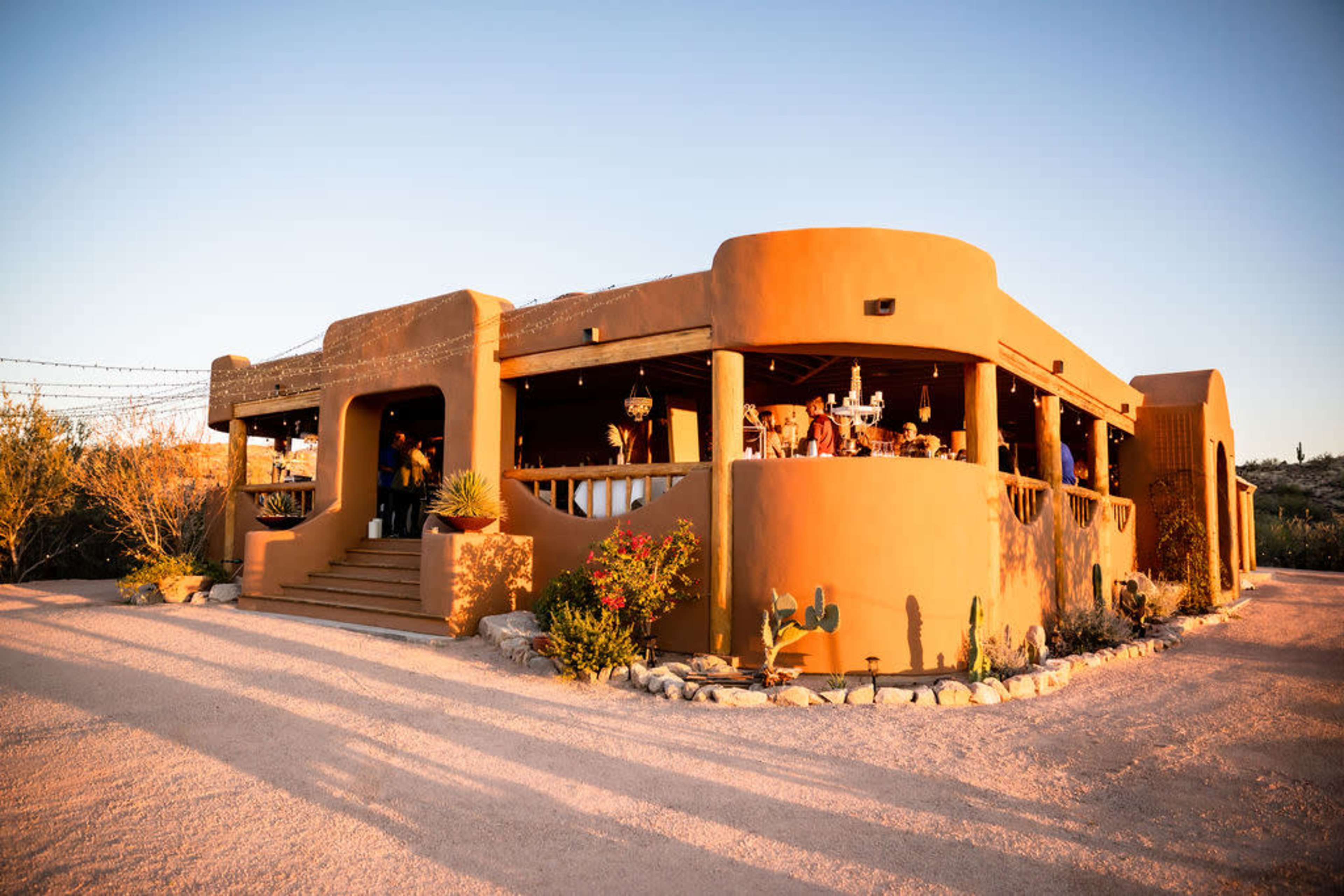 A tan adobe-style building with a rounded roof and decorative accents is surrounded by desert landscaping and bathed in warm sunlight.