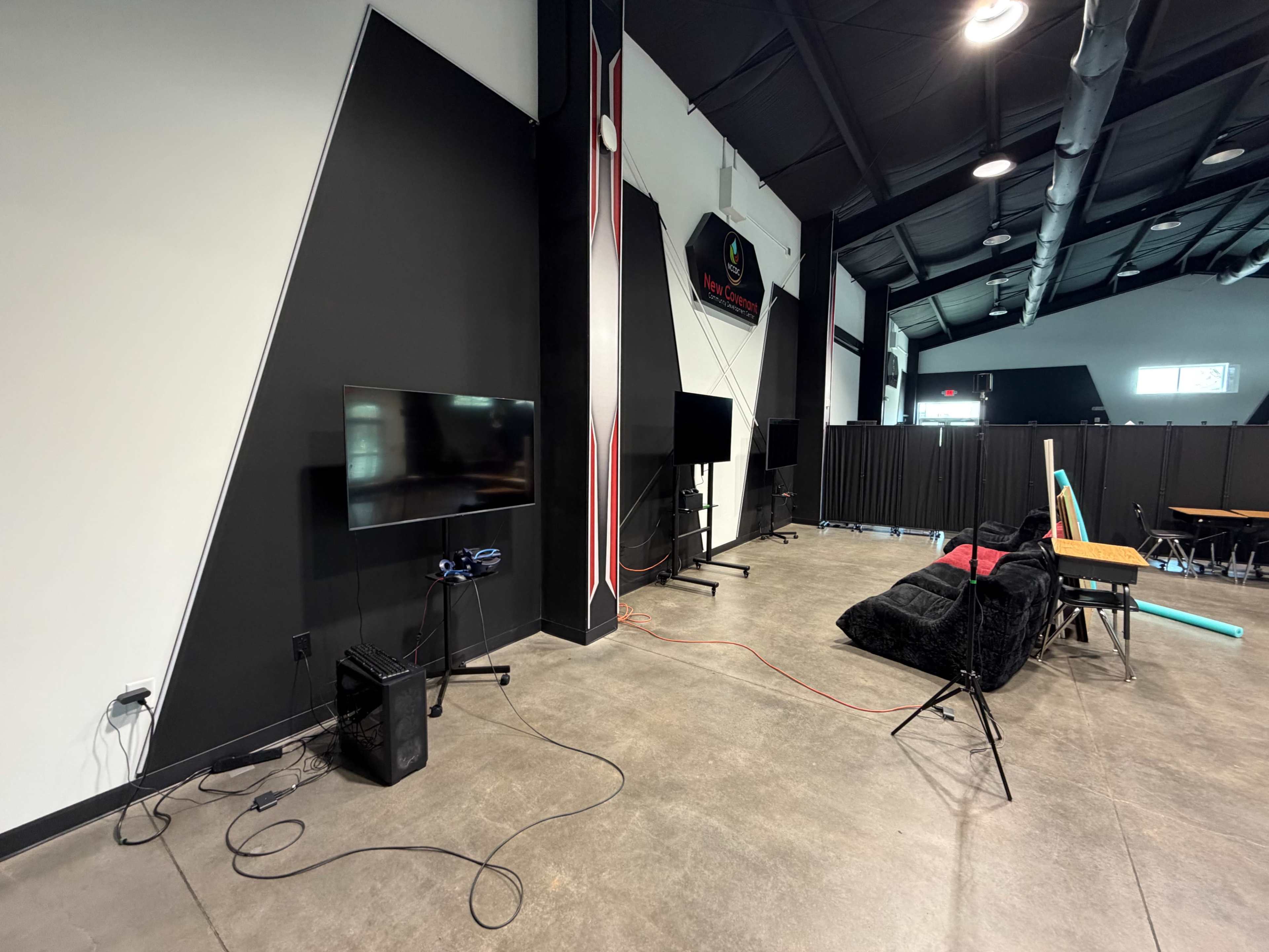 The image shows a corner of a room featuring a wall-mounted television, a stand-alone monitor, and a black bean bag chair on a gray floor.