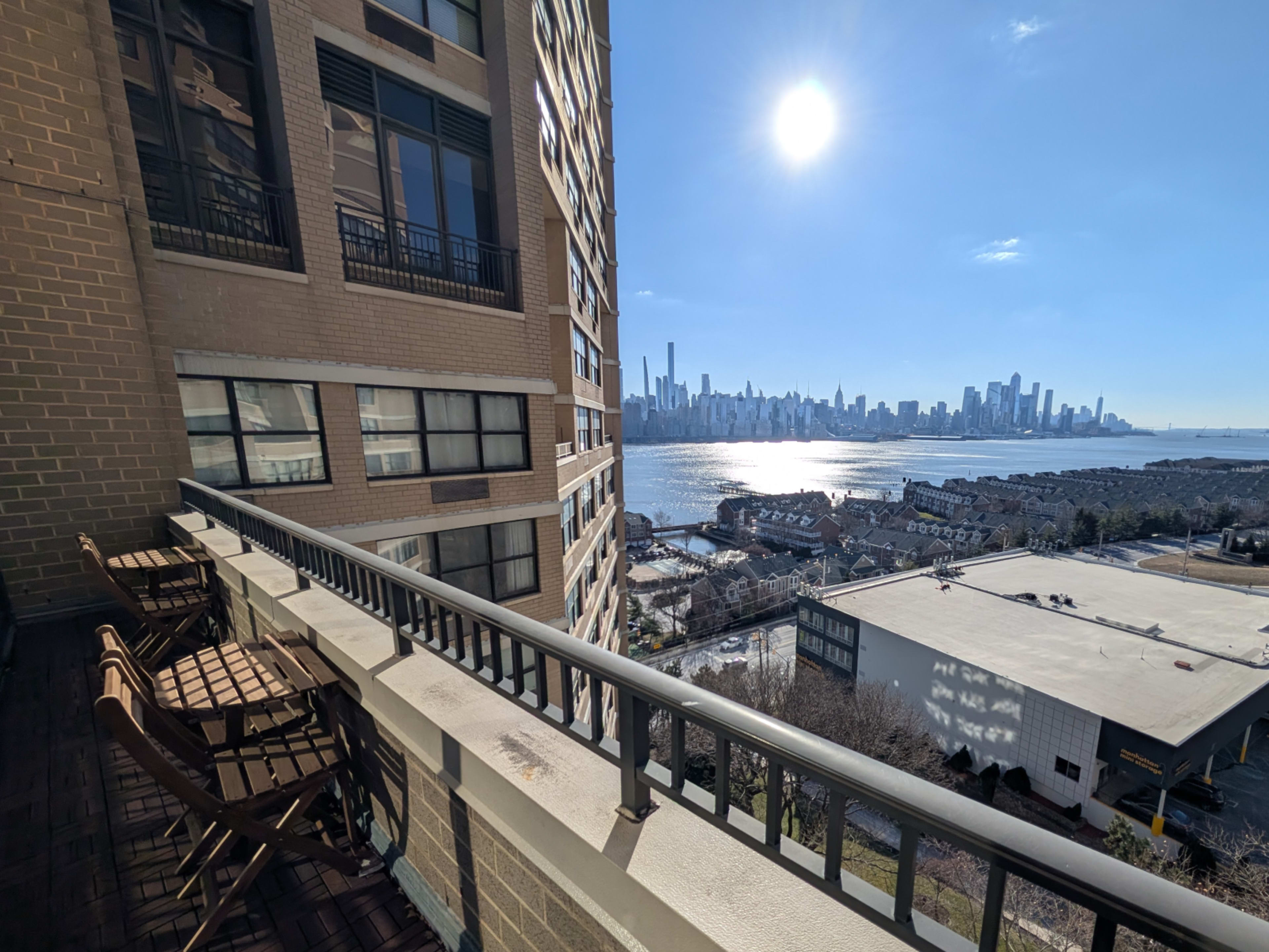 A balcony overlooks a river with a city skyline and bright sun in the background.