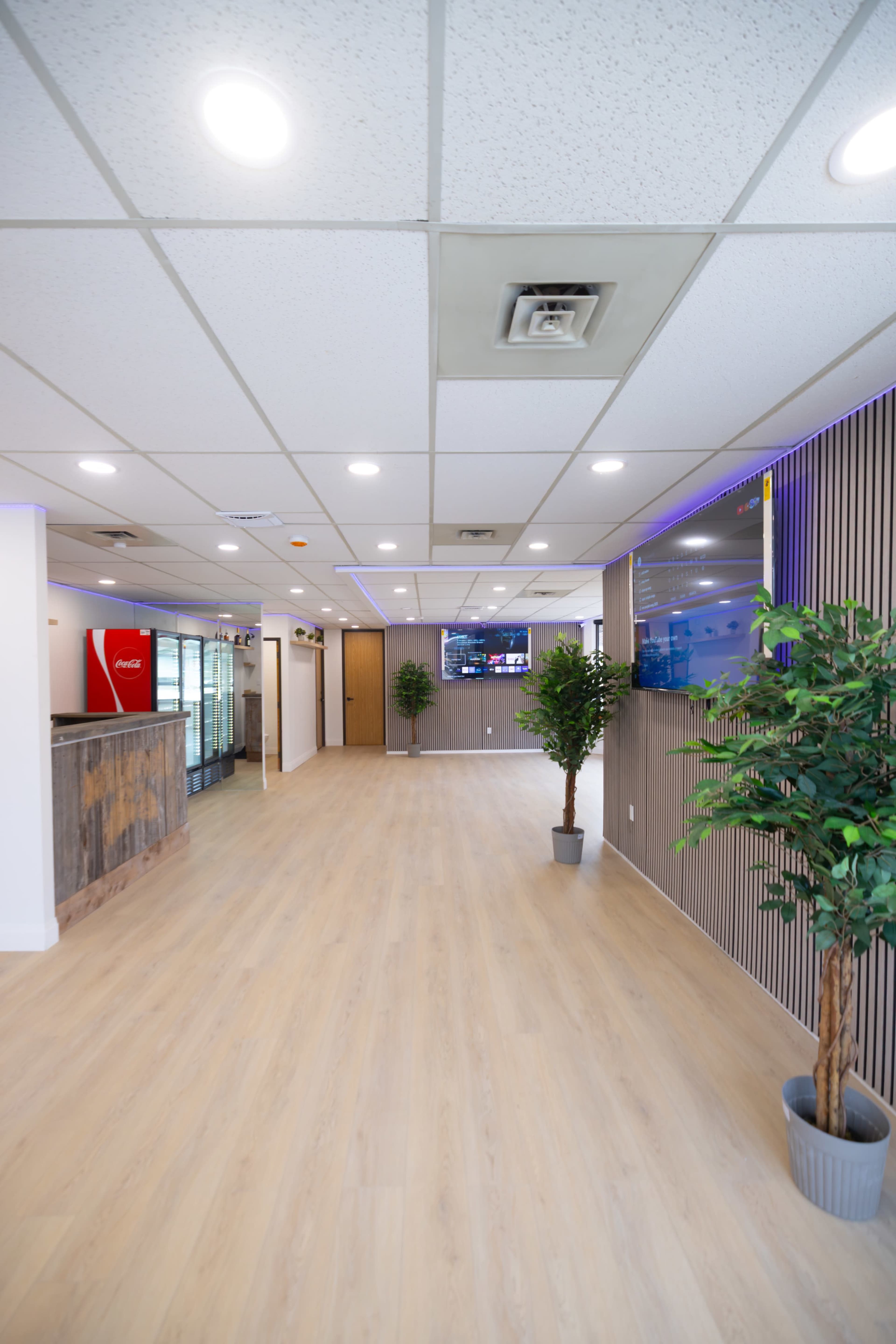 The image shows a modern, spacious office reception area with light wooden flooring, potted plants, and multiple screens displaying information on the walls.