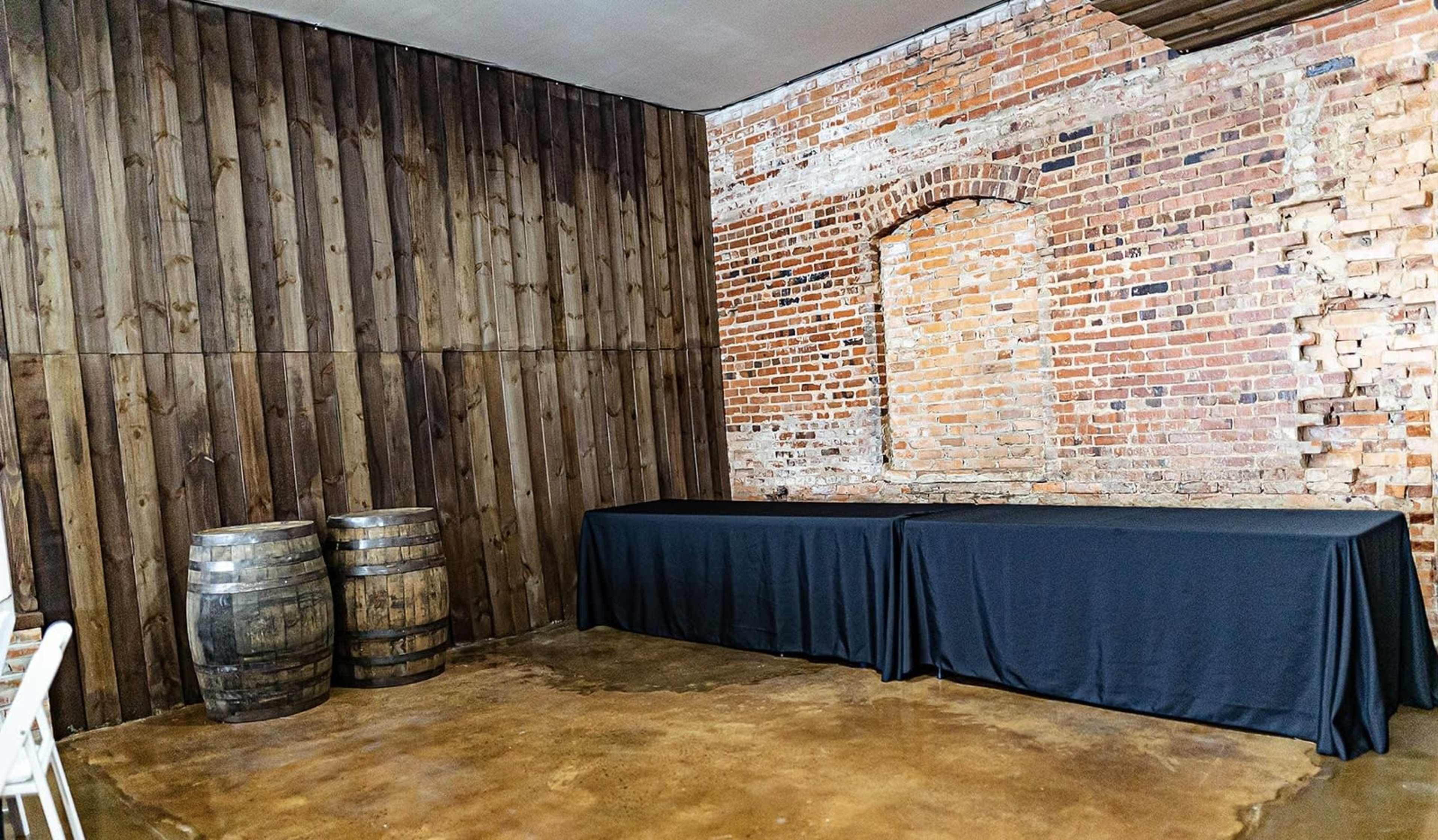 A room with a wooden wall and exposed brick, featuring two barrels and two tables covered with black tablecloths.
