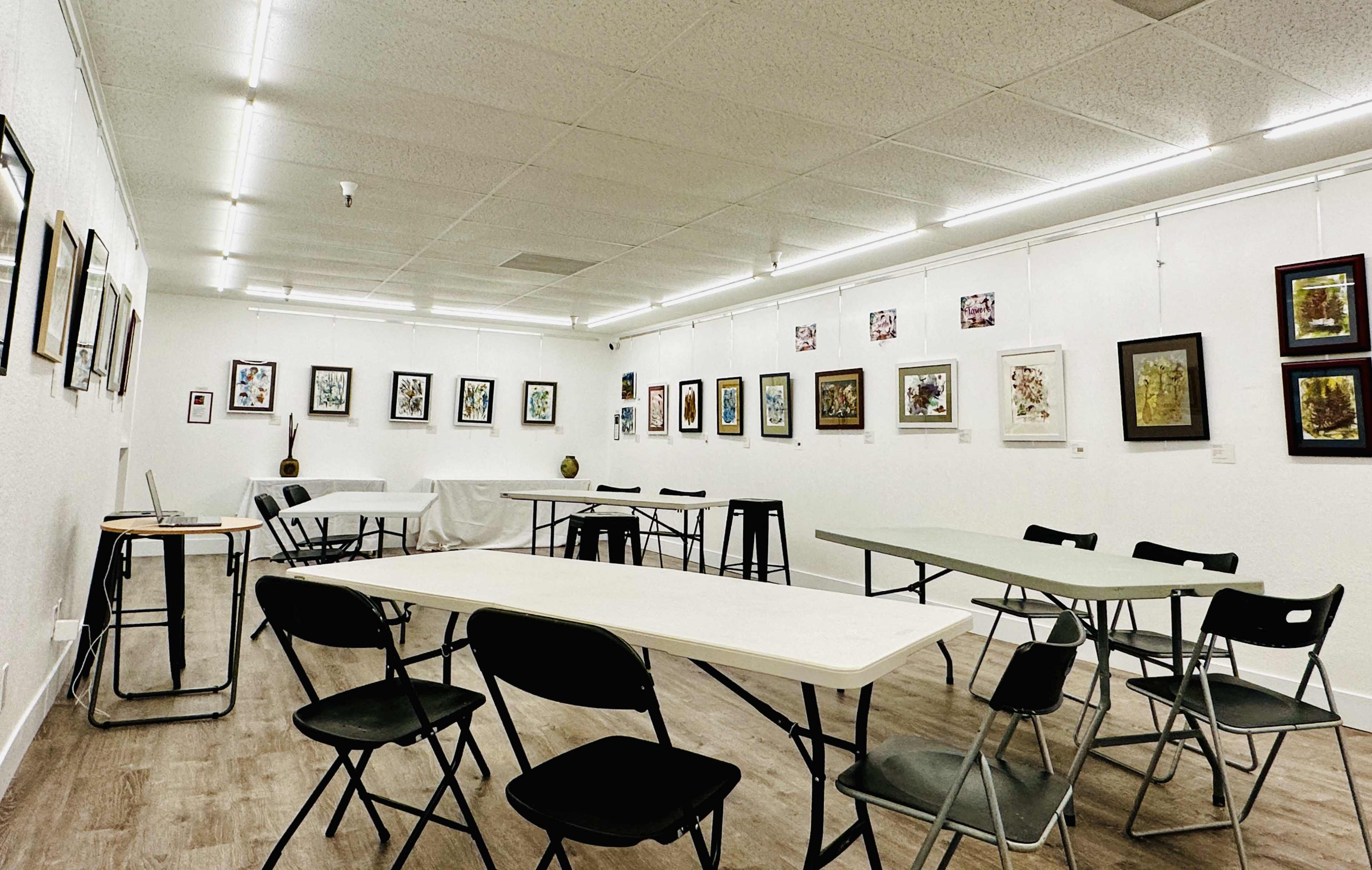 An art gallery with tables and chairs arranged around the room, and various framed artworks hanging on the walls.