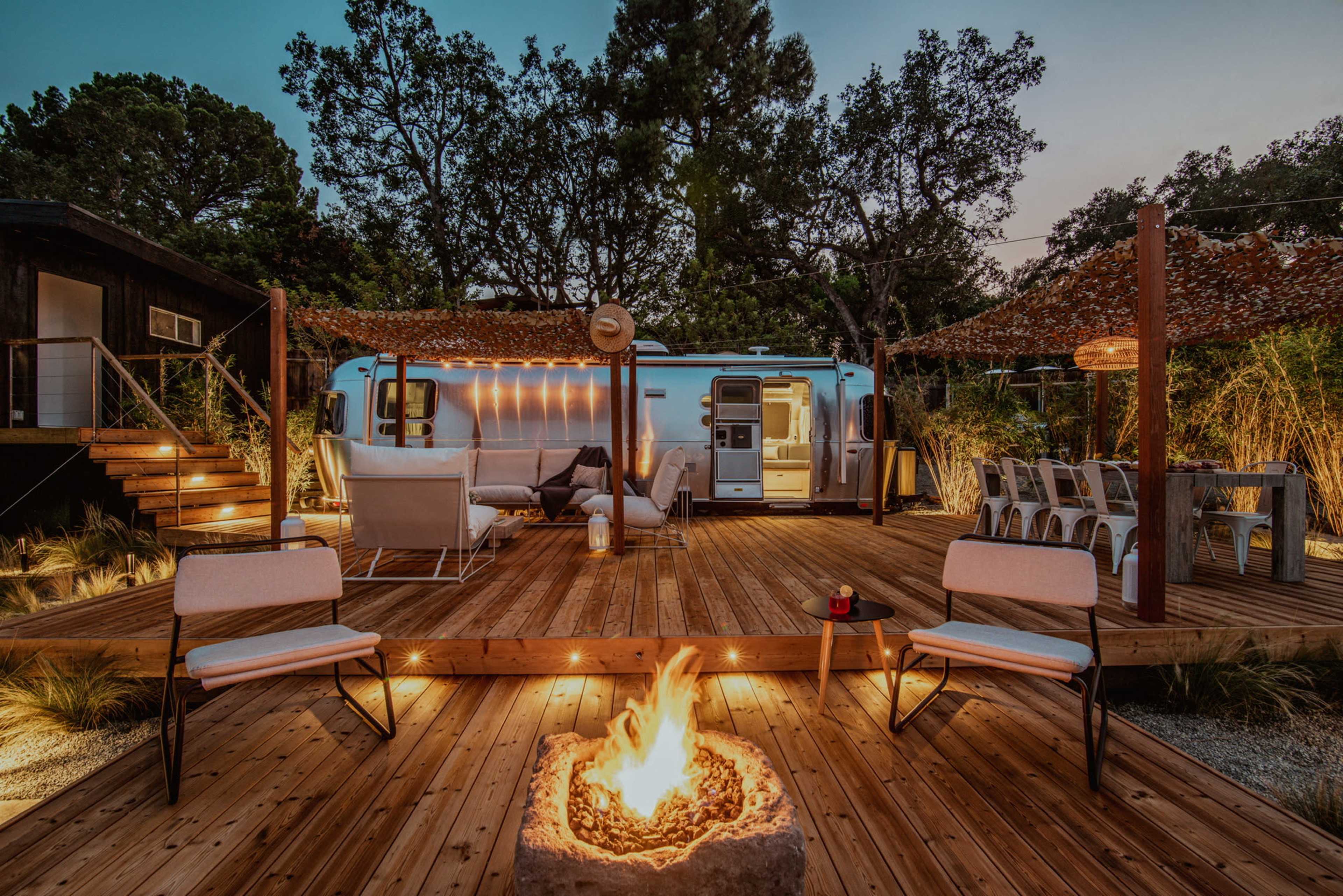 A cozy outdoor space features a fire pit surrounded by chairs, a deck with comfortable seating, and an Airstream trailer illuminated by string lights amidst trees.