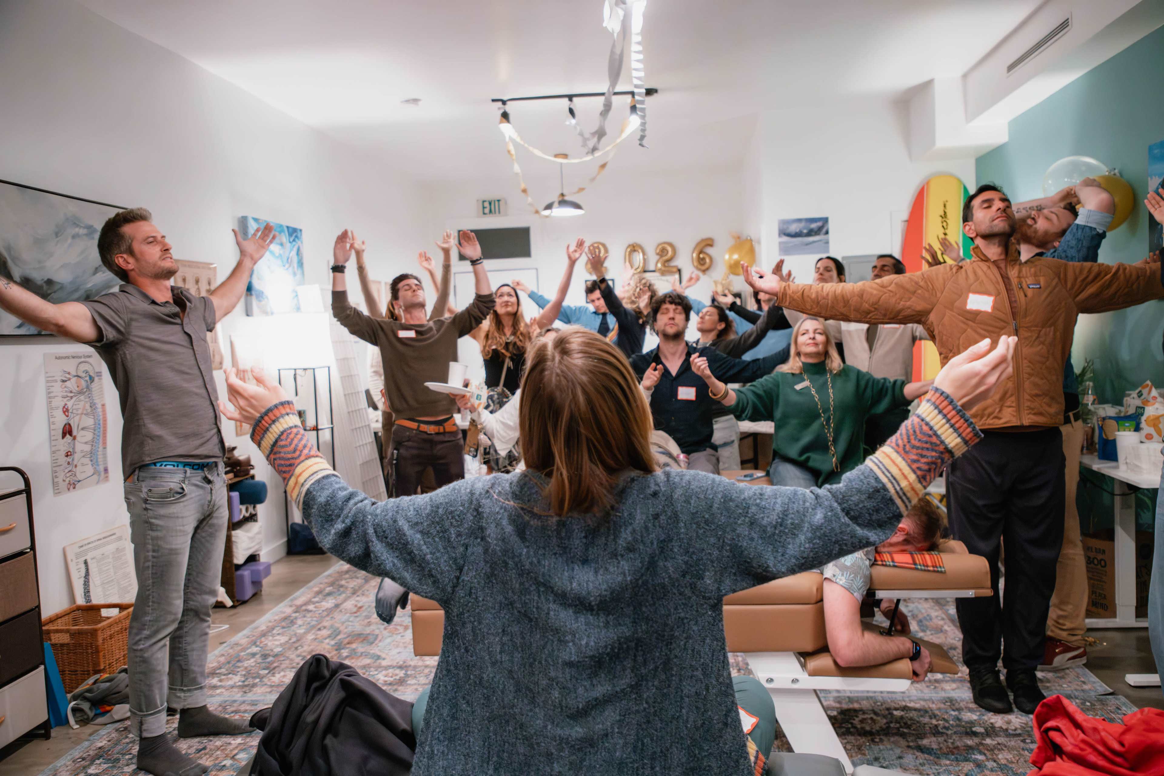 A group of people is participating in a group activity, standing with their arms raised in a room decorated with various items and a banner that reads "2026."