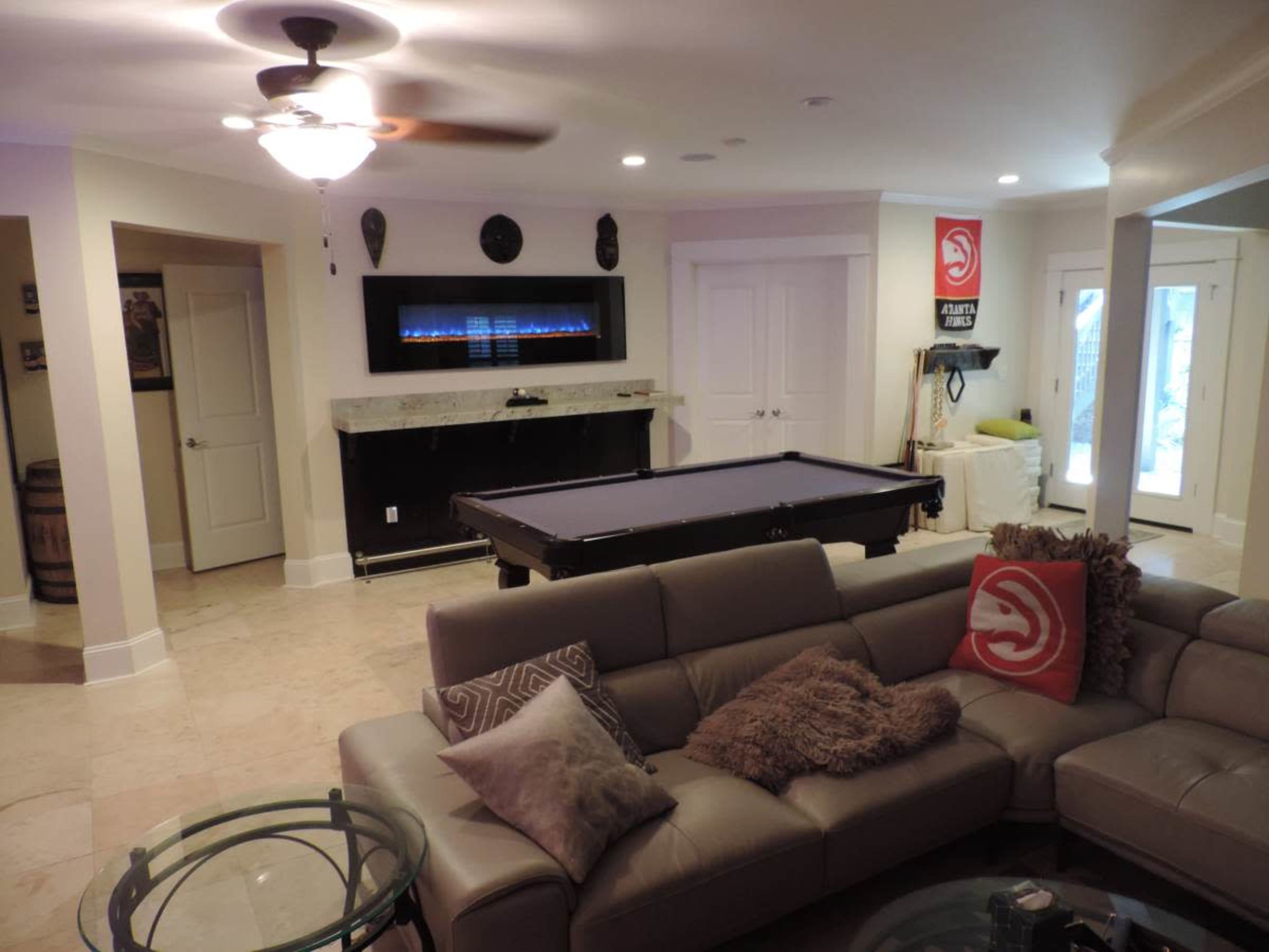 The image shows a modern game room featuring a pool table, a cozy sectional sofa, and an electric fireplace along with a sliding glass door leading to the outdoors.