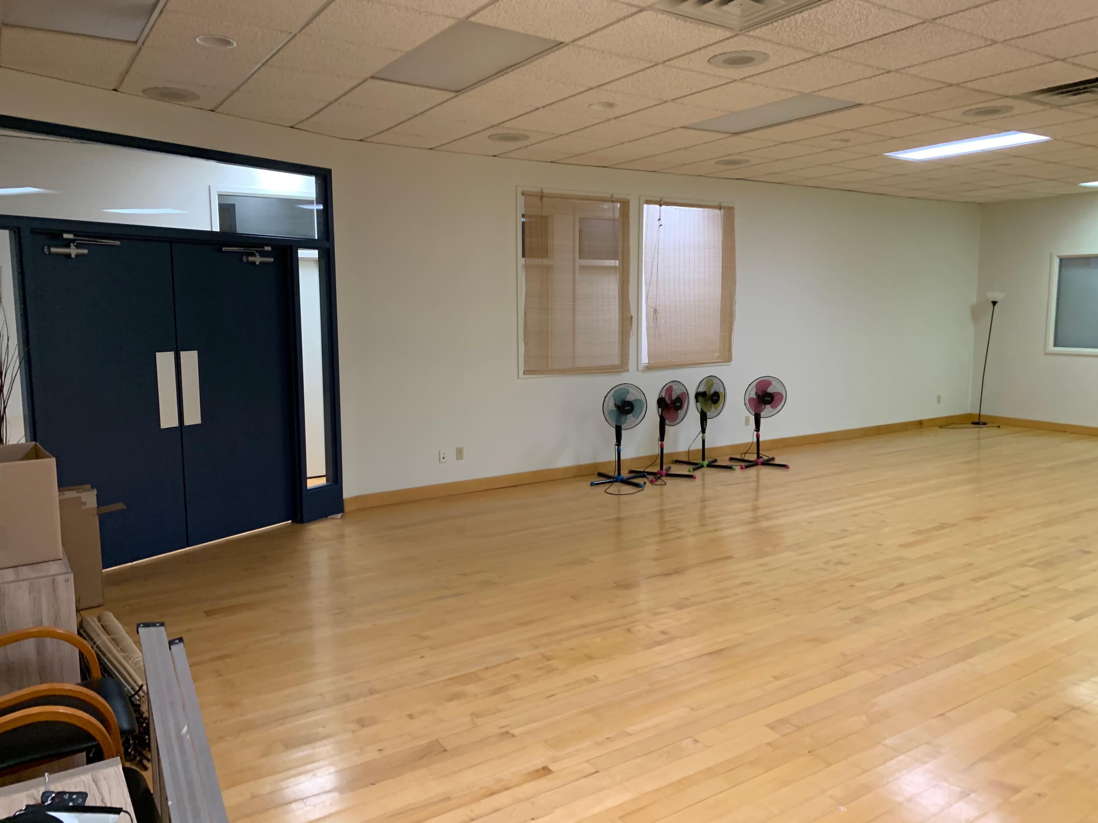An empty room with wooden flooring, two doors on the left, a window with blinds, and four standing fans lined up against the wall.