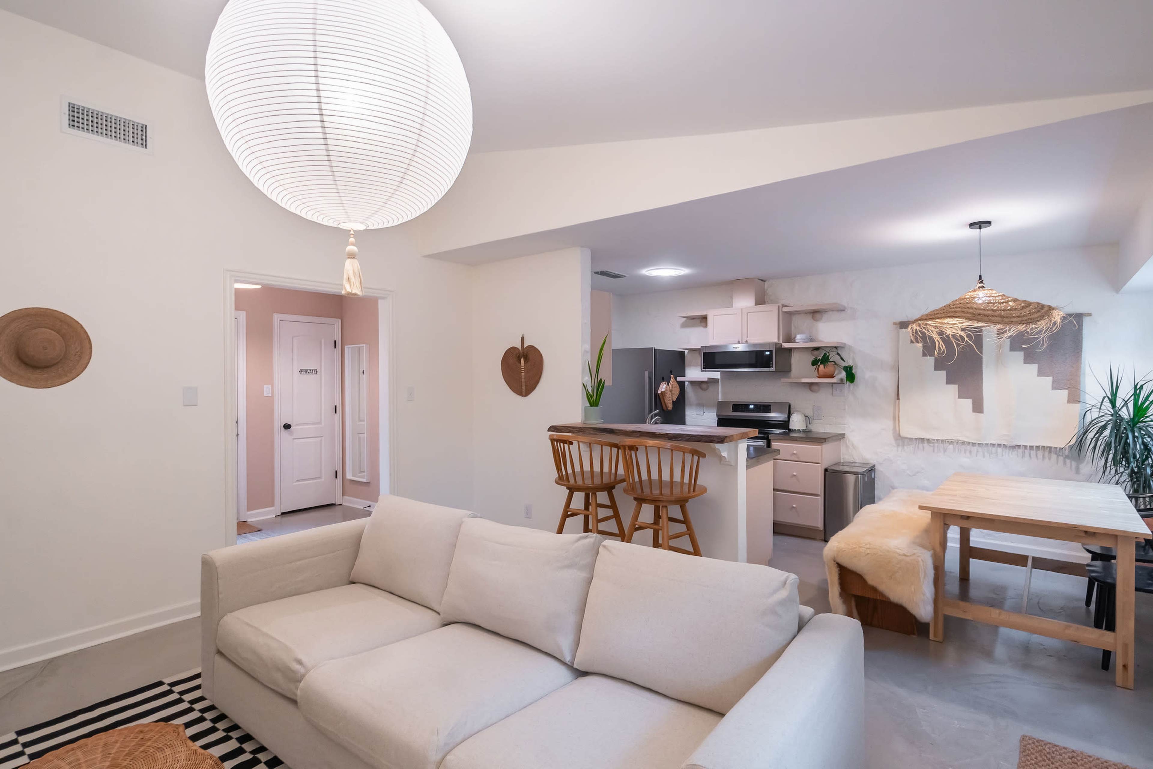 A modern living space featuring a light-colored sofa, a kitchen with bar stools, and a dining area with a wooden table.