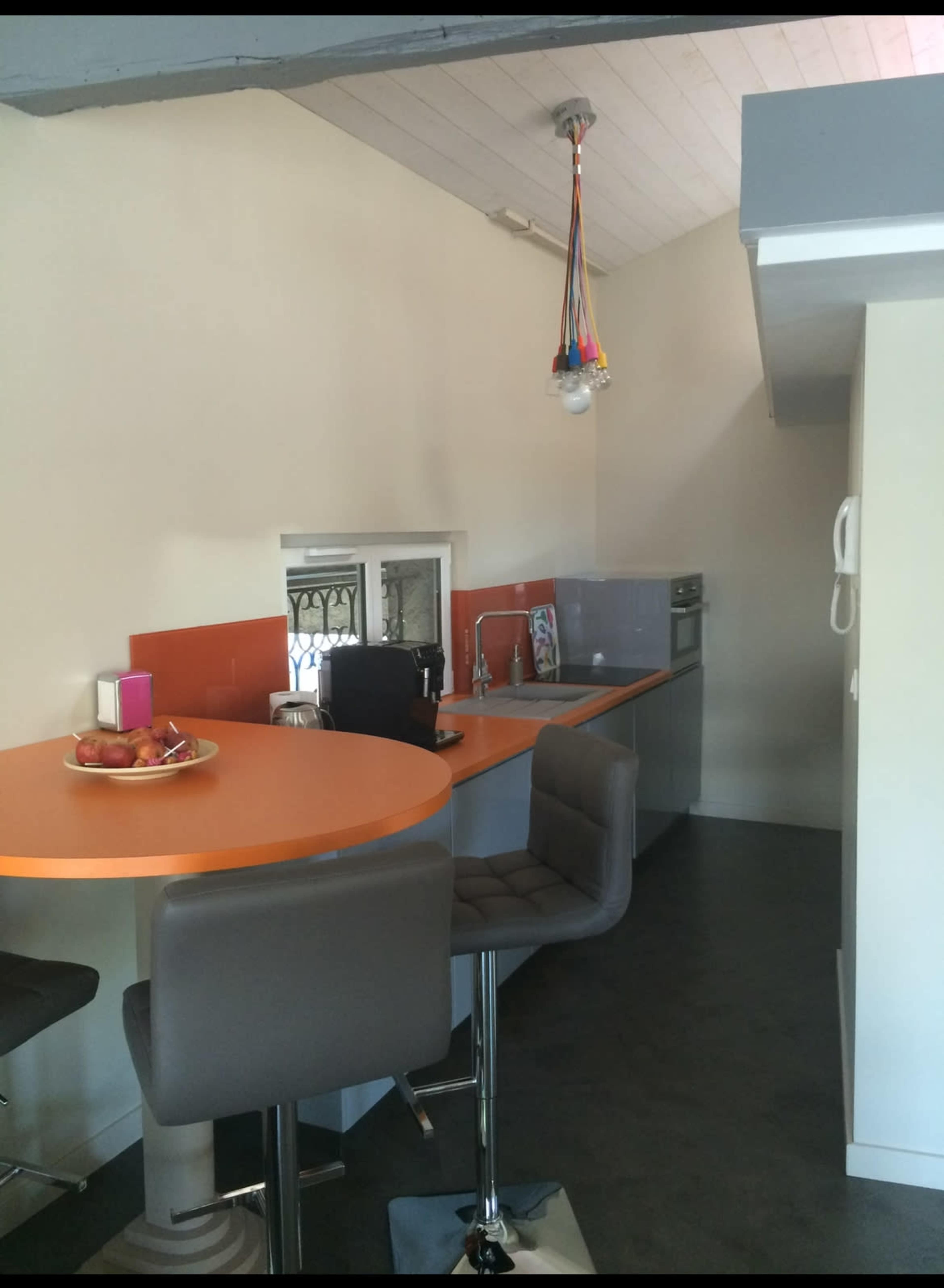 The image shows a modern kitchen space featuring a bar with four stools, an orange countertop, and stainless steel appliances against a light-colored wall.