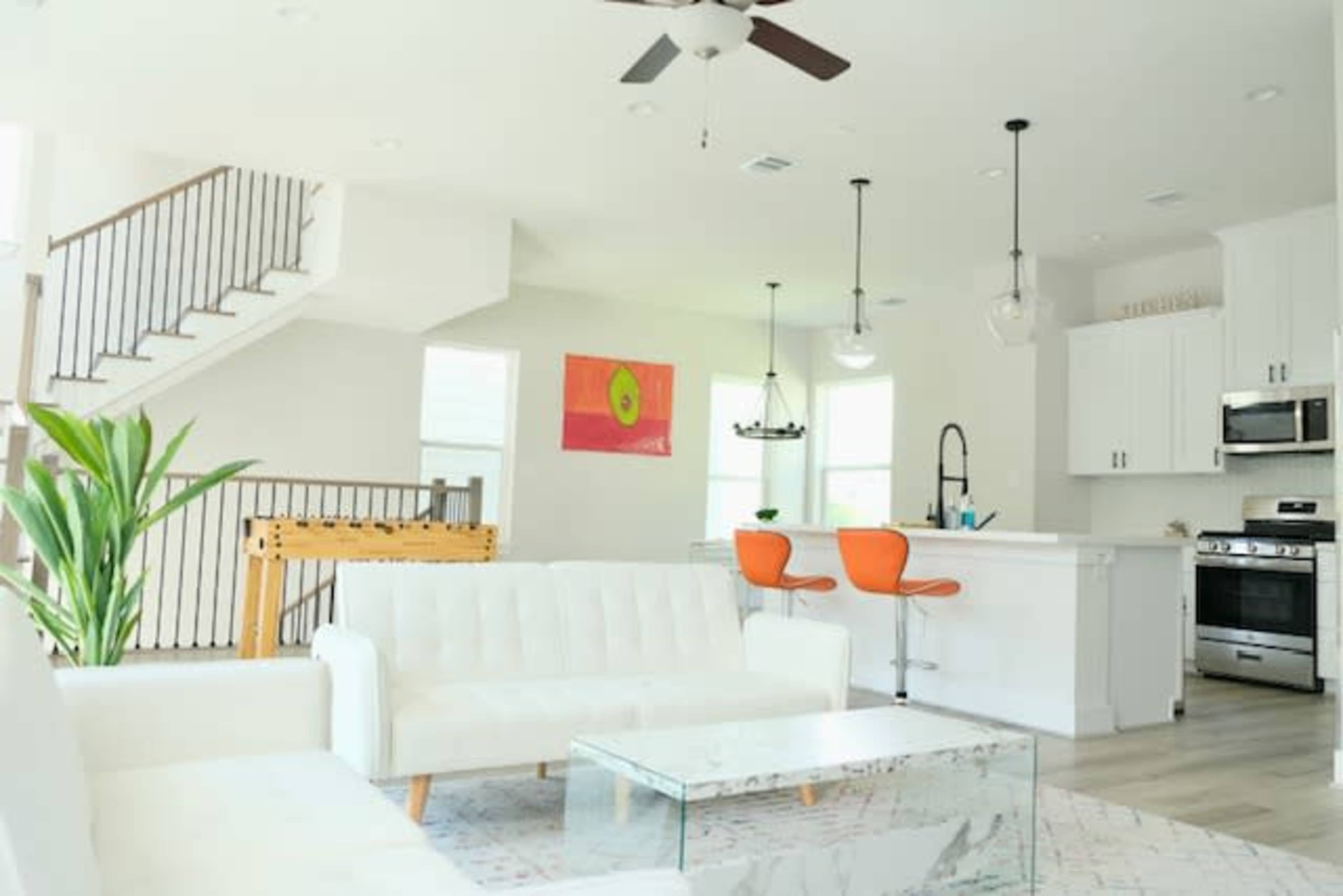 The image shows a modern living area with a white sofa, a glass coffee table, and an adjacent kitchen featuring orange bar stools and stainless steel appliances.