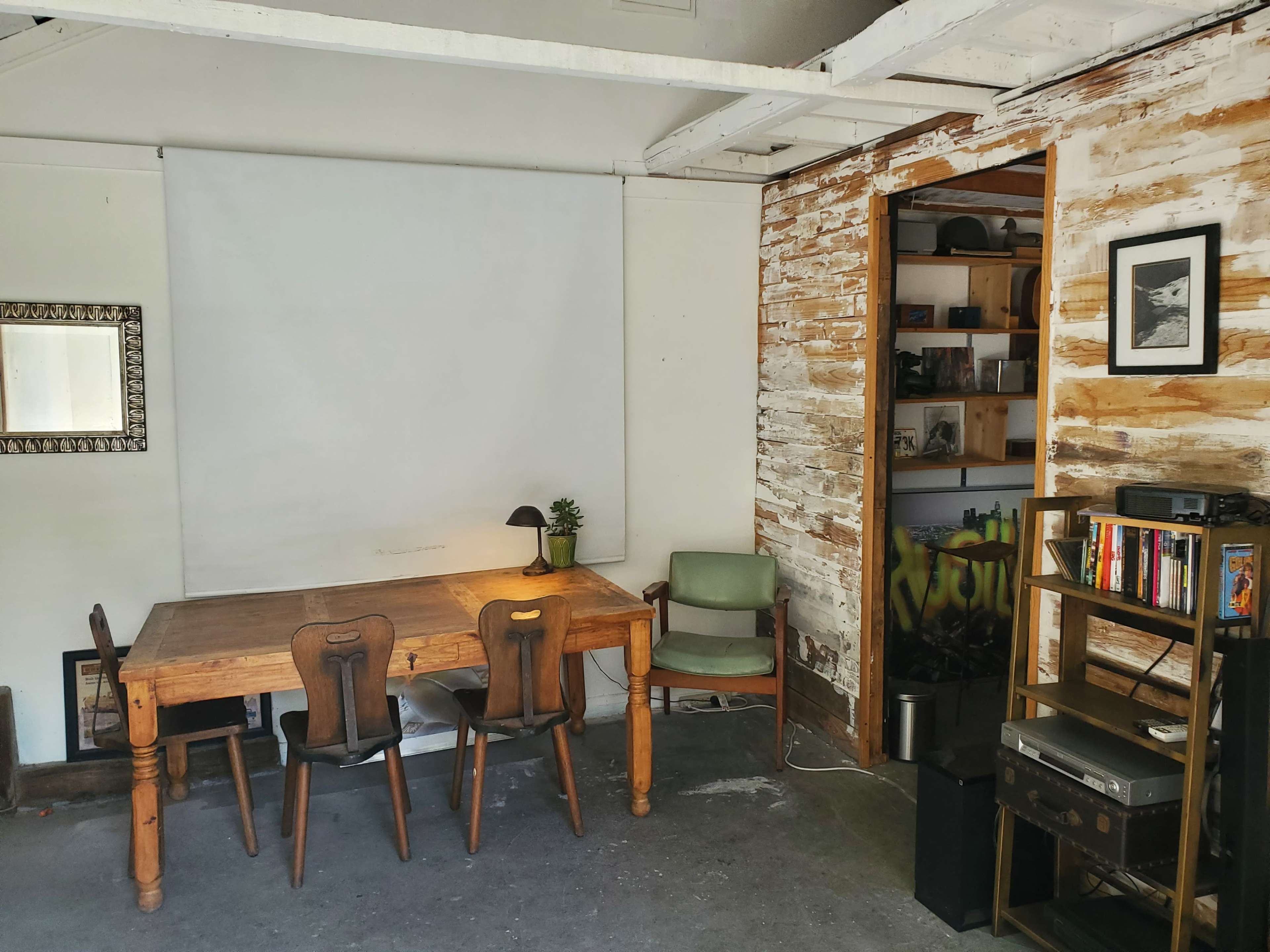 The image shows a simple room with a wooden table and three chairs, a lamp on the table, and a bookshelf in a corner against a wall with an uneven wooden texture.
