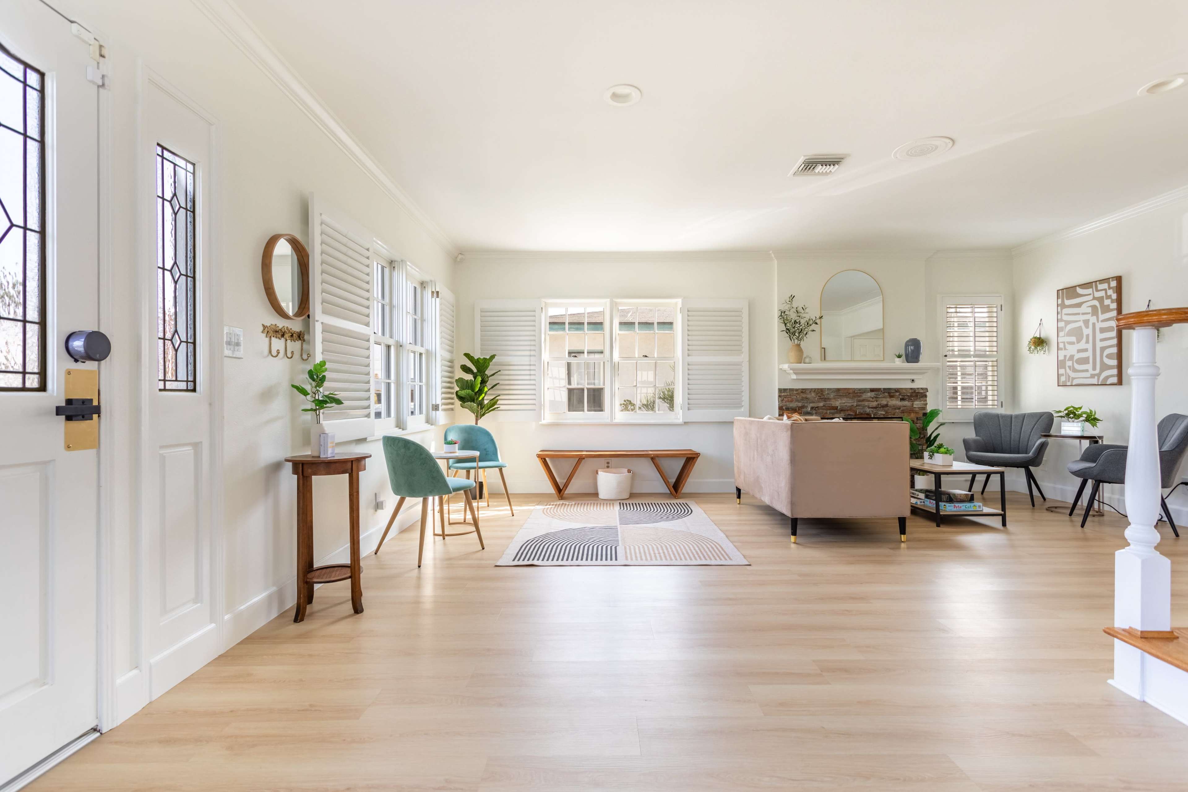 The image shows a spacious living room with light-colored walls, wooden flooring, a mix of seating options, and large windows allowing natural light to enter.