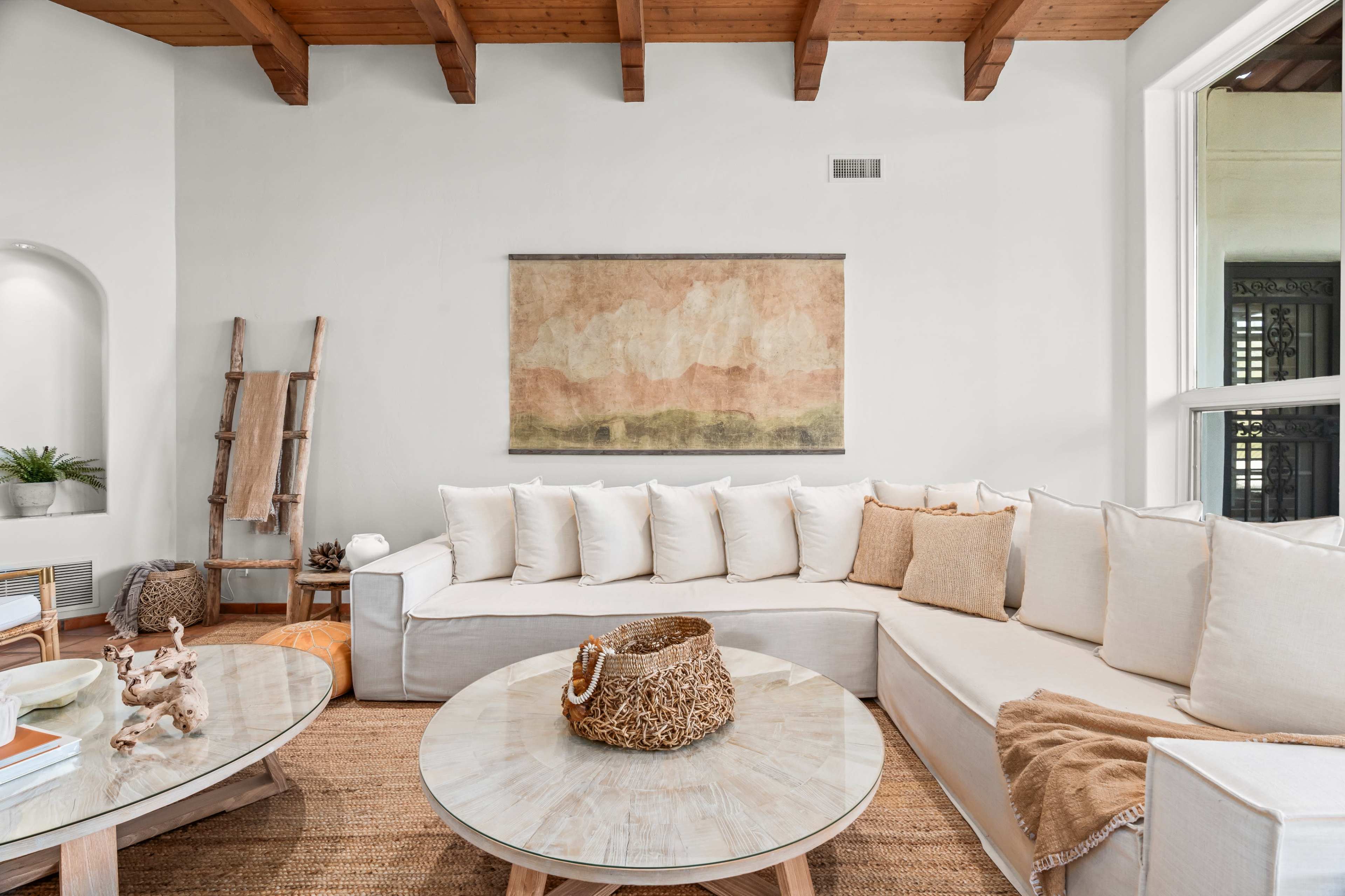 A spacious living room features a large corner couch with white cushions, a circular coffee table, and a textured area rug beneath wooden beams.
