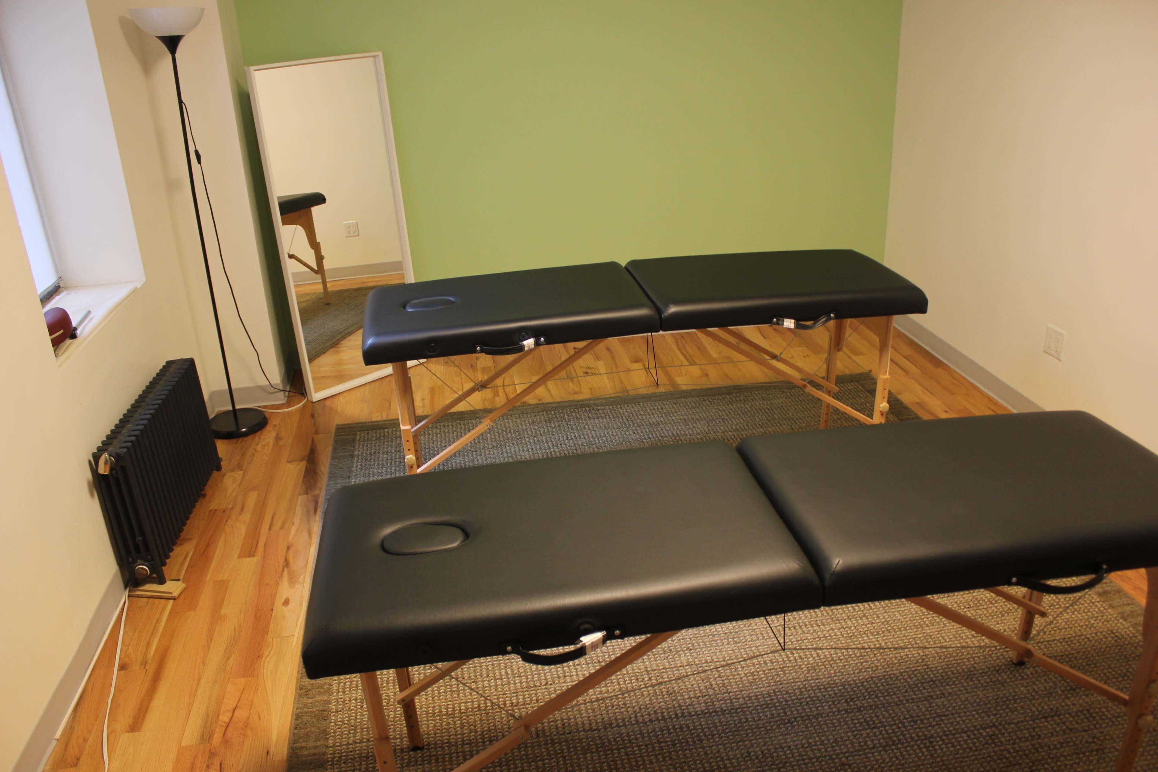 A massage room with two black massage tables, a floor lamp, and a mirror against a green wall.
