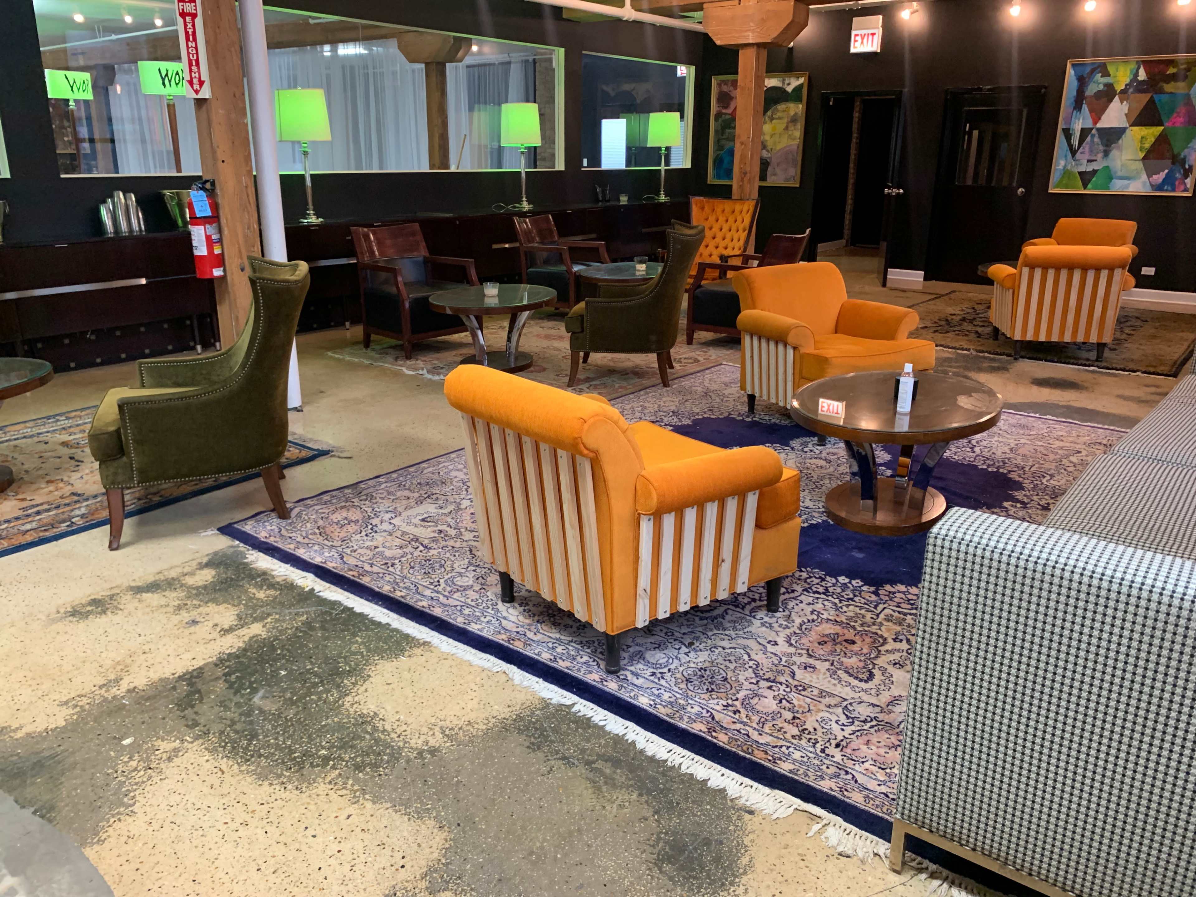 The scene shows a lounge area with several chairs and tables set on a patterned rug, surrounded by green lamps and artwork on the walls.