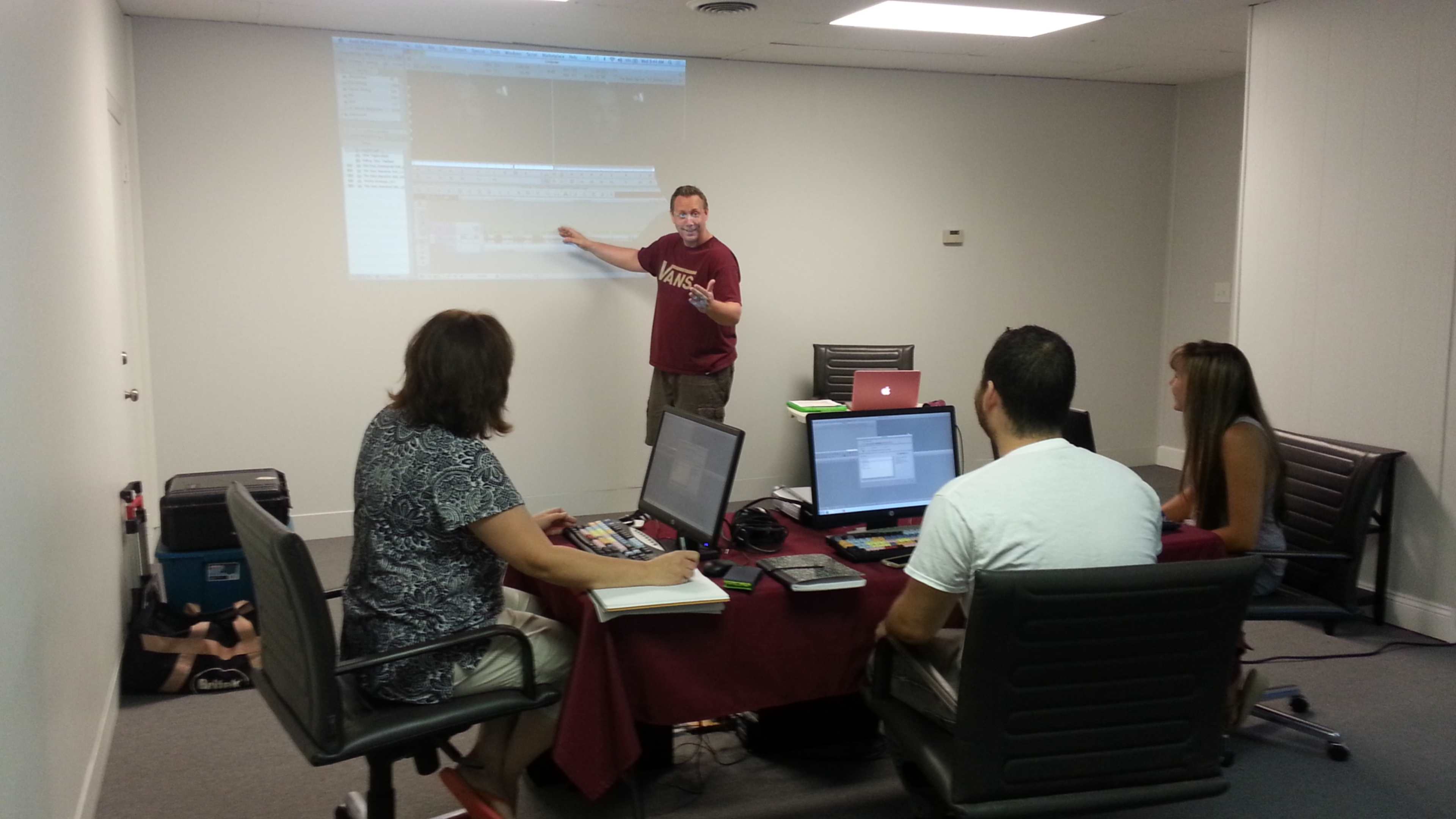 A man stands by a large screen while presenting to a group seated around a table with laptops and materials during a workshop.
