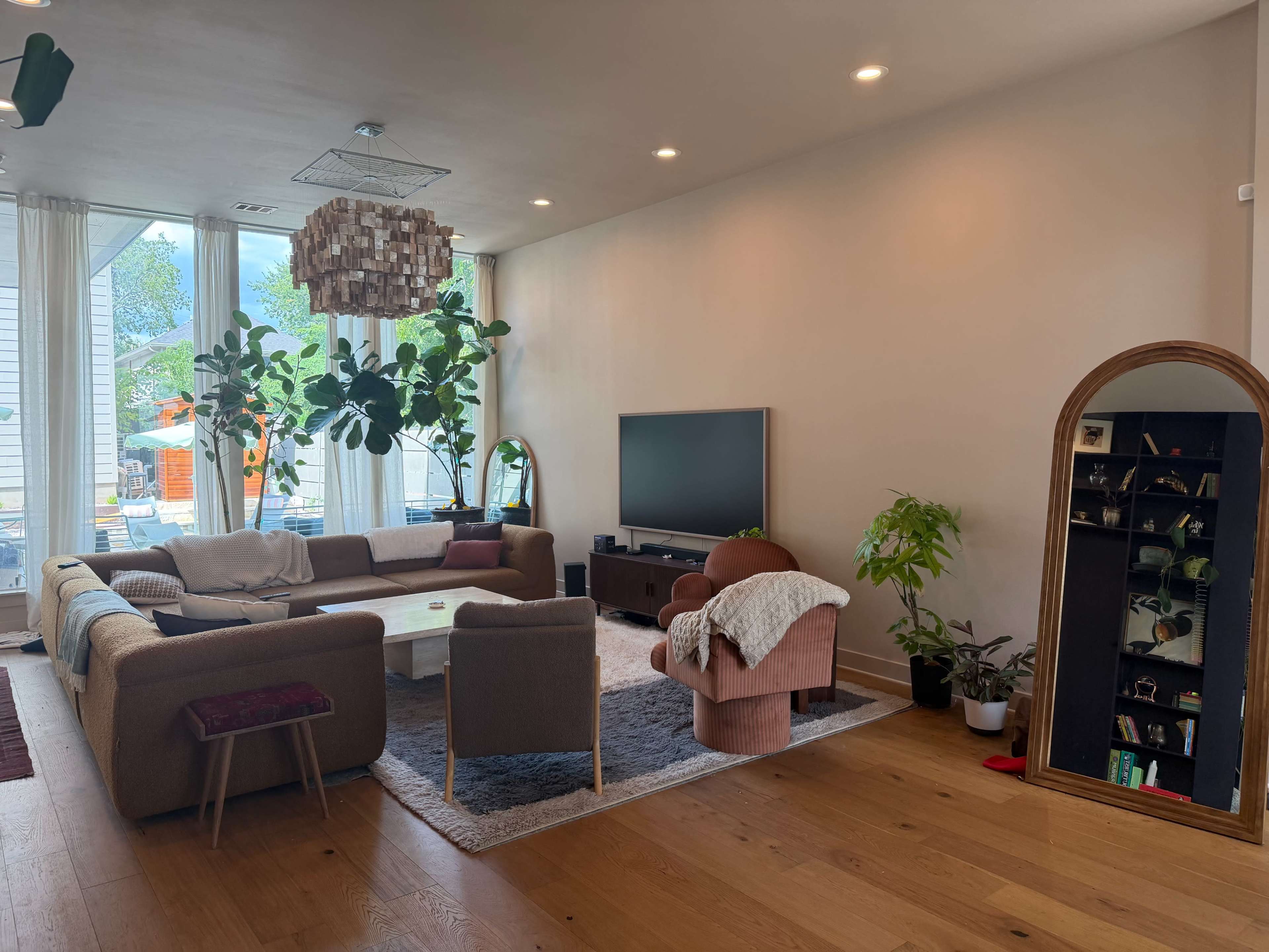 The image depicts a modern living room featuring a large sectional sofa, a television on a low stand, lush indoor plants, and a large mirror against a wall.