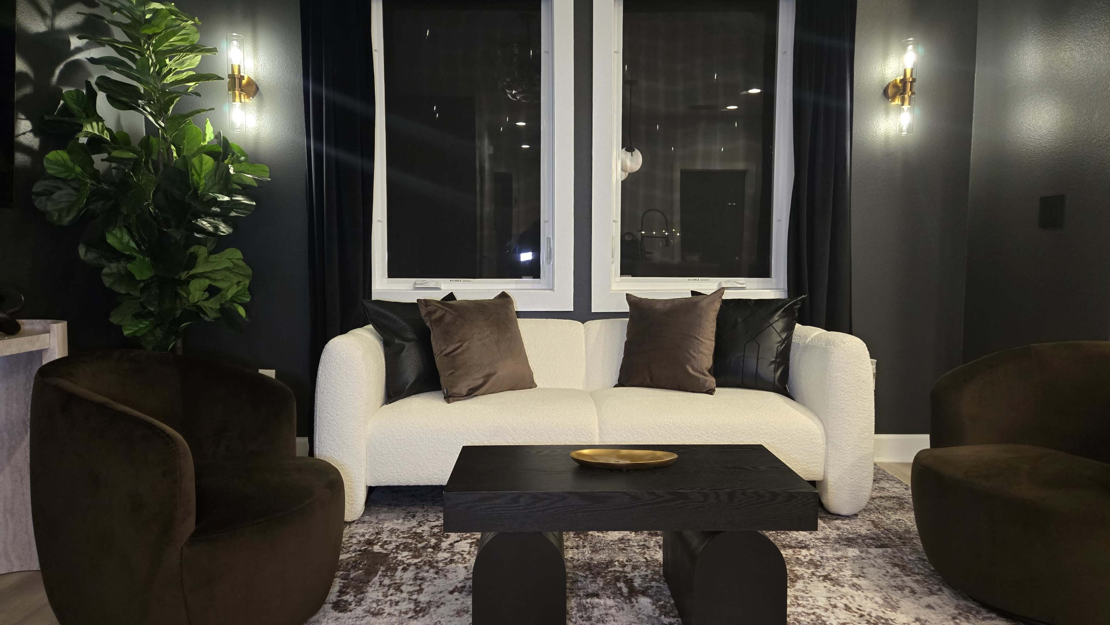 The image shows a modern living room with a white, textured sofa adorned with dark pillows, two brown upholstered chairs, a dark wooden coffee table, and large windows covered by dark curtains.