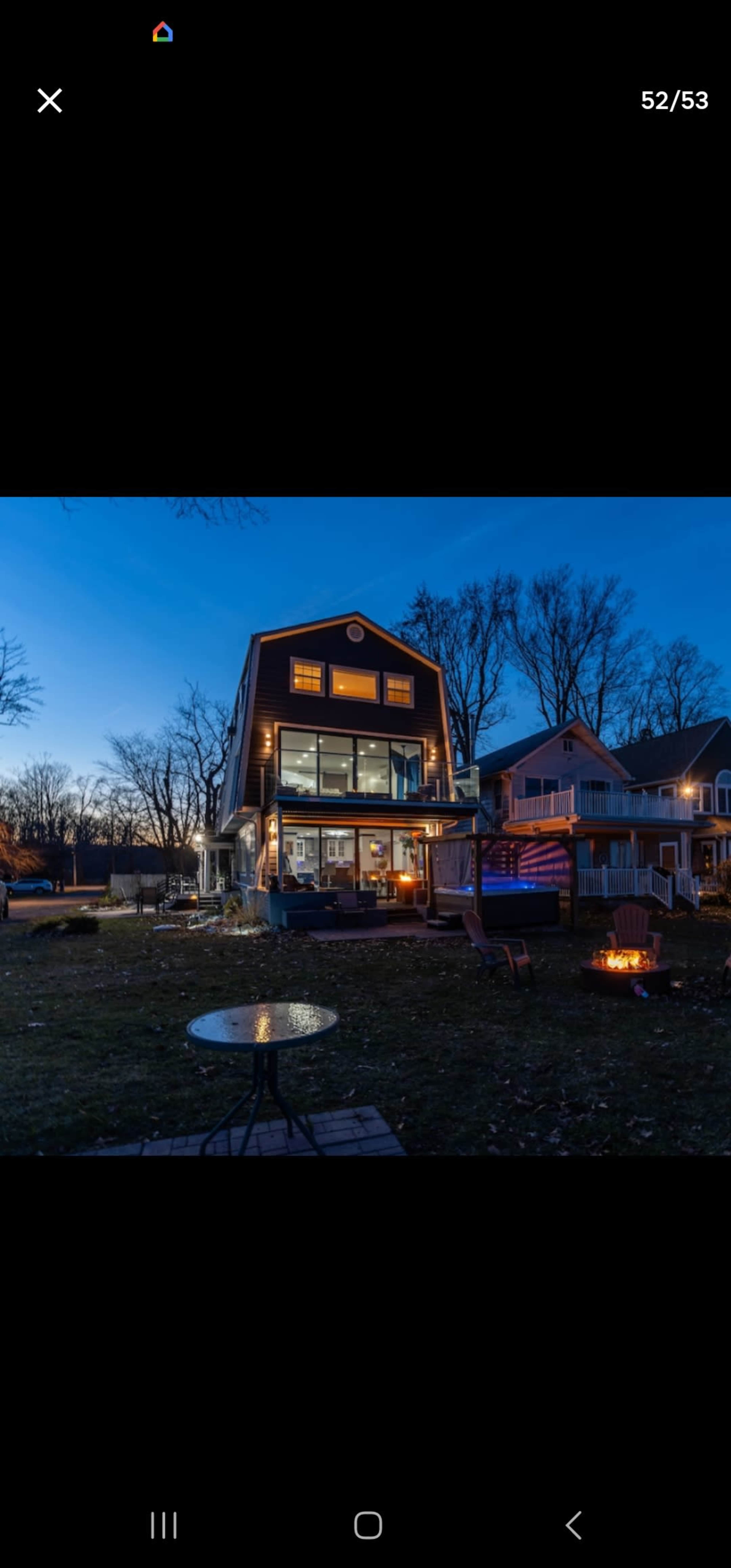 A modern two-story house with large windows and an outdoor seating area, set against a twilight sky.