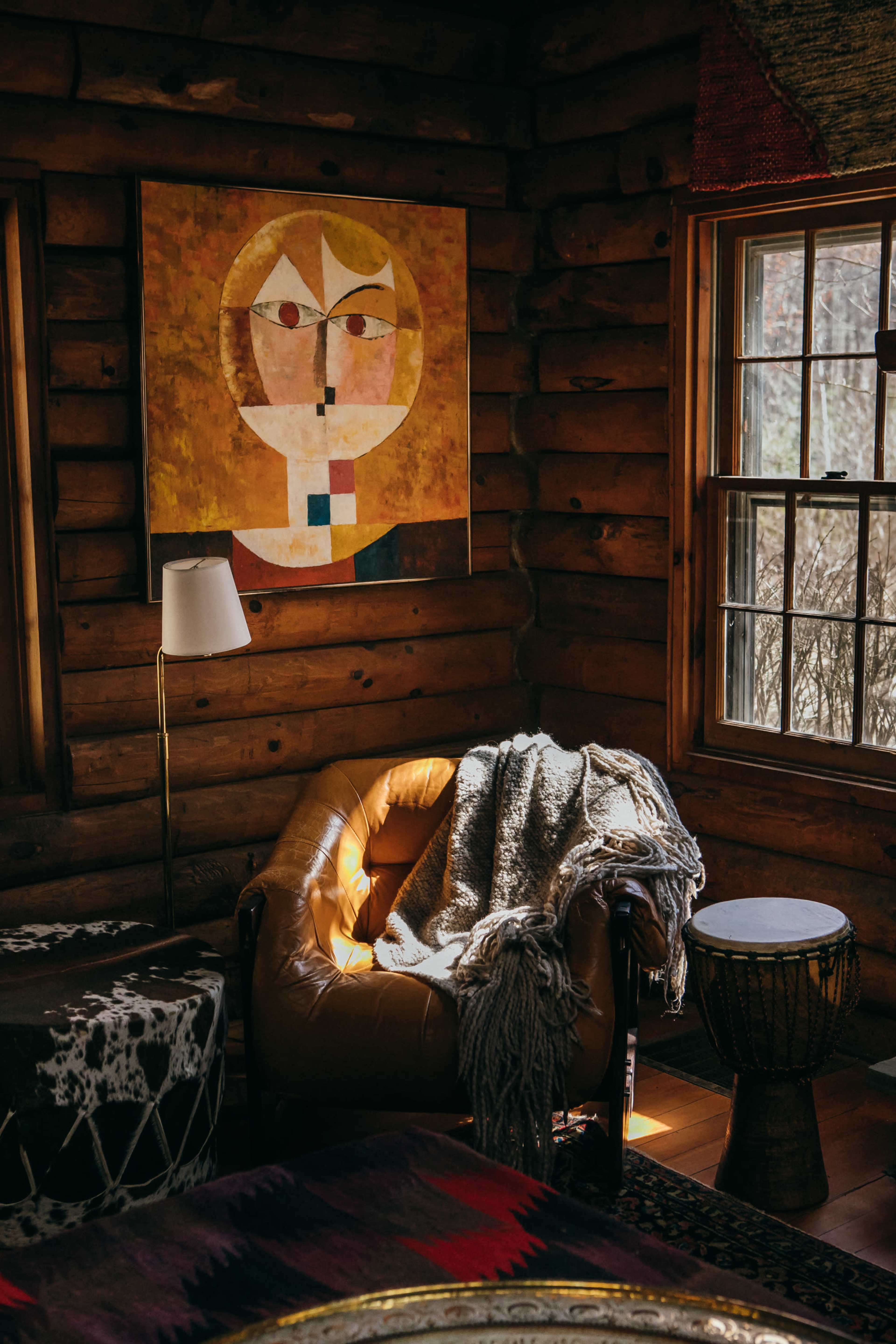 A leather armchair with a gray blanket draped over it is positioned next to a wooden side table and a lamp beneath a colorful abstract painting on a log cabin wall.