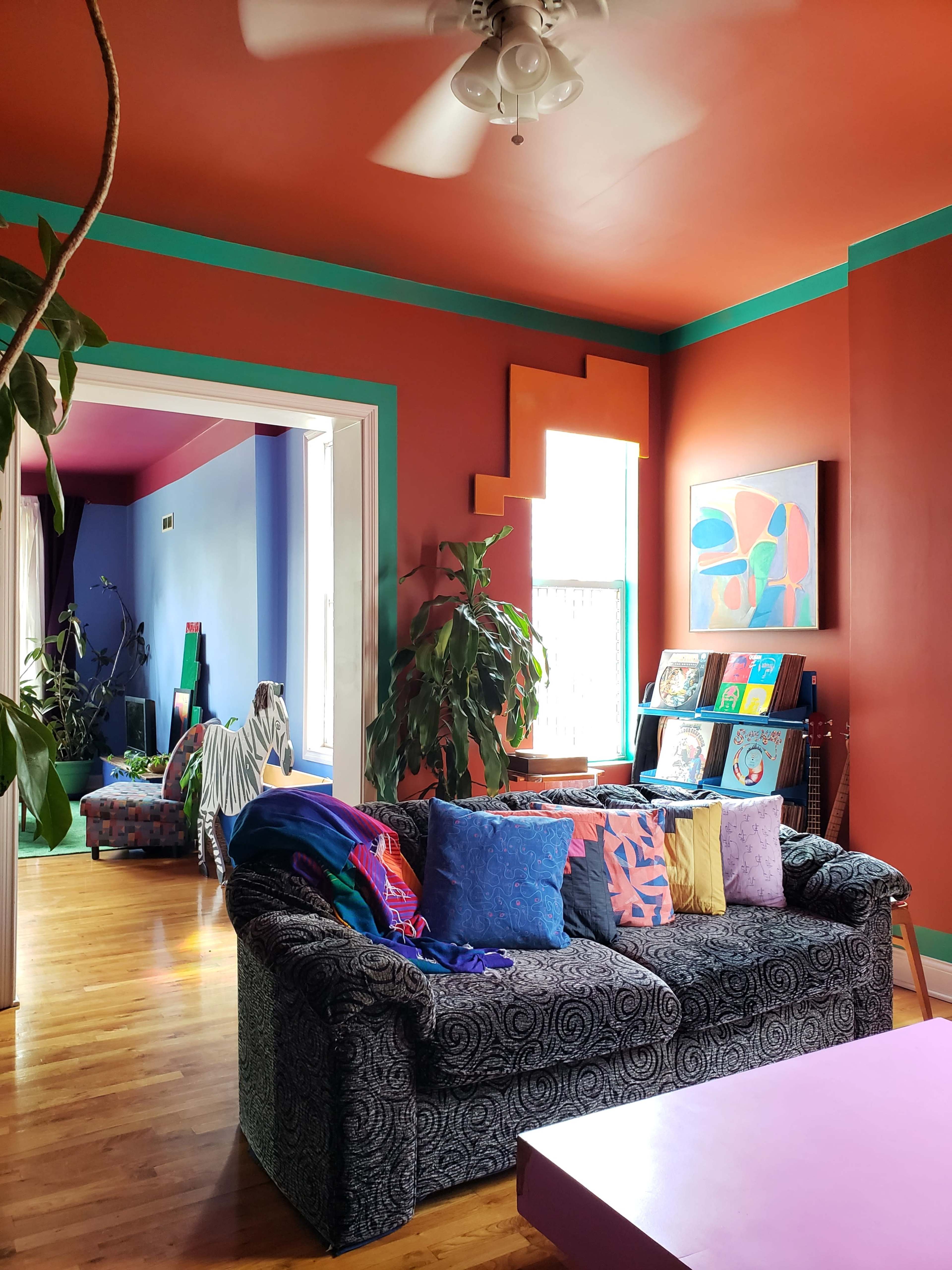 A living room featuring a dark patterned sofa adorned with colorful pillows, vibrant orange walls, and an adjoining blue room visible through an open entryway.