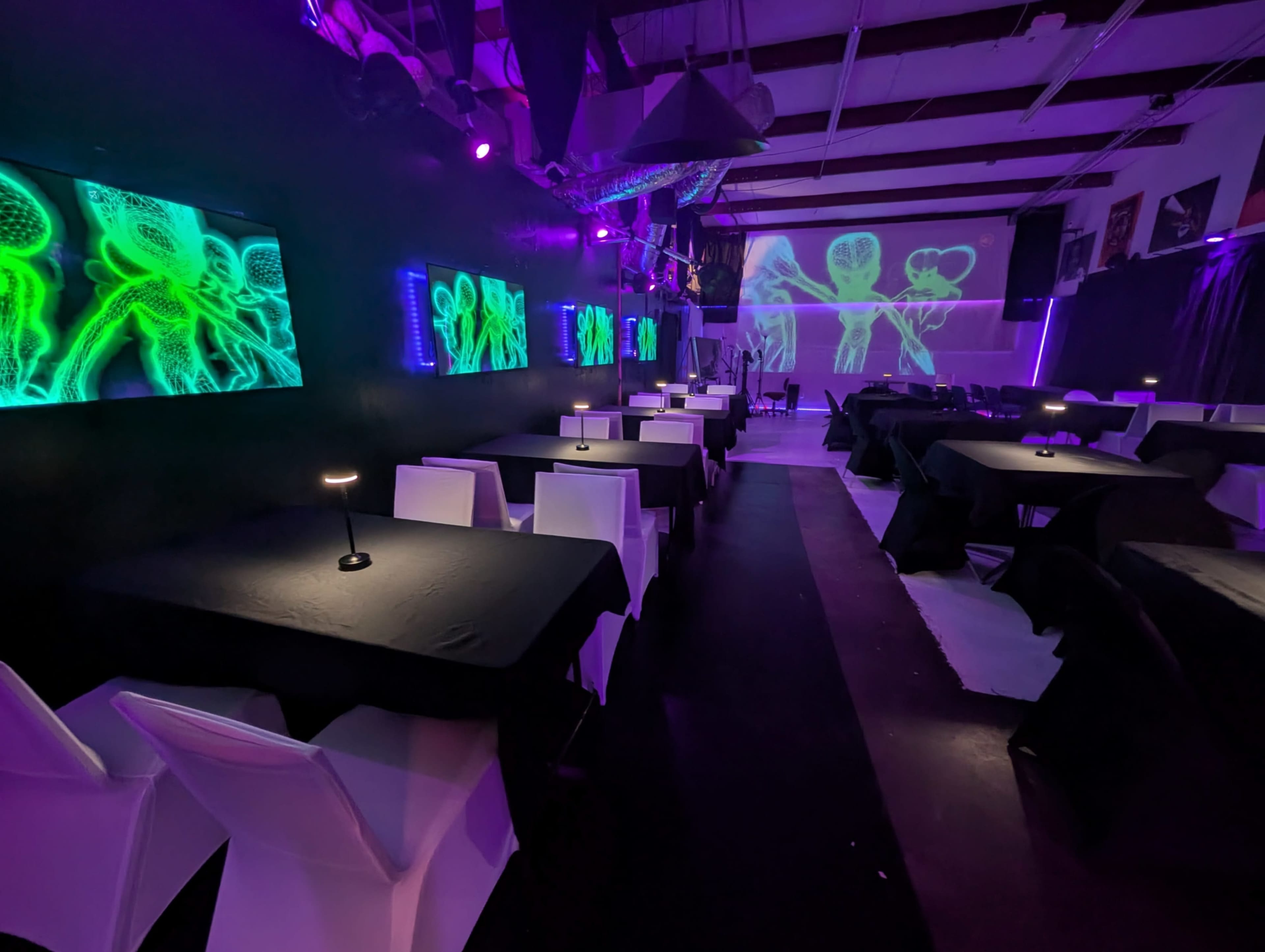 The interior of a dimly lit venue features black tables set for dining, illuminated by purple lights and adorned with neon artwork on the walls.