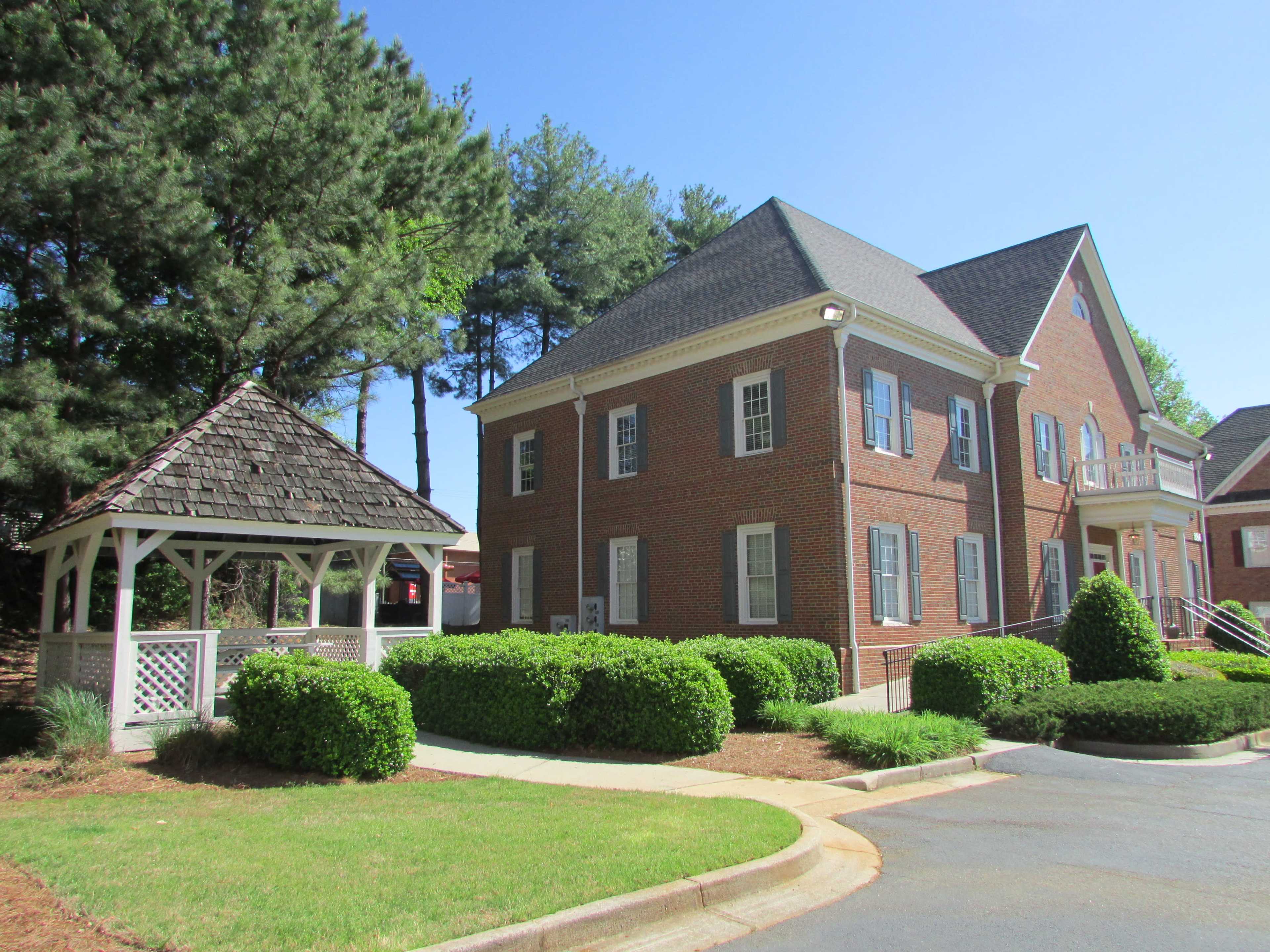 A brick building next to a gazebo surrounded by neatly trimmed shrubs and trees.
