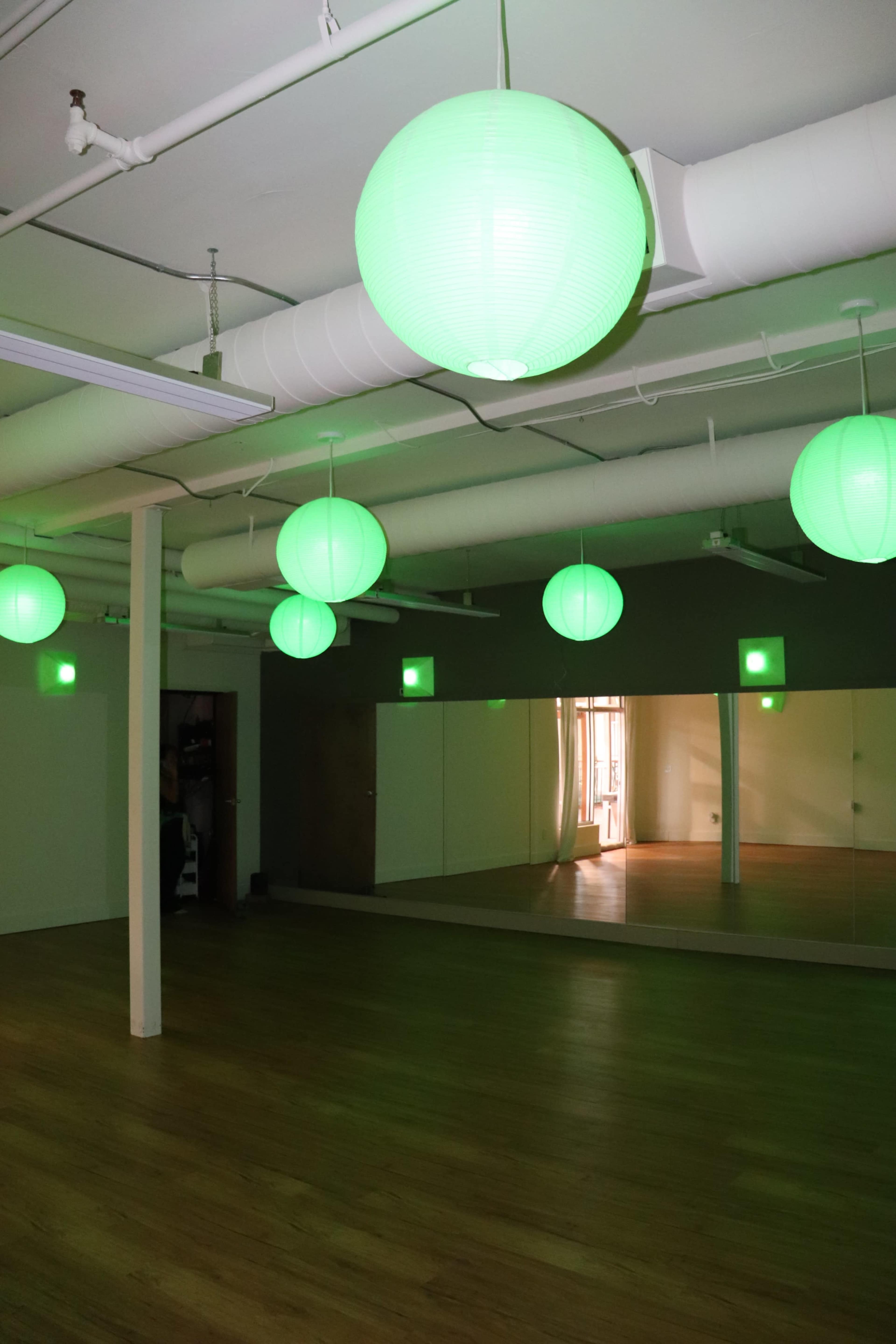 The image shows a spacious room with wooden flooring, featuring green paper lanterns hanging from the ceiling and a large mirror on one wall.