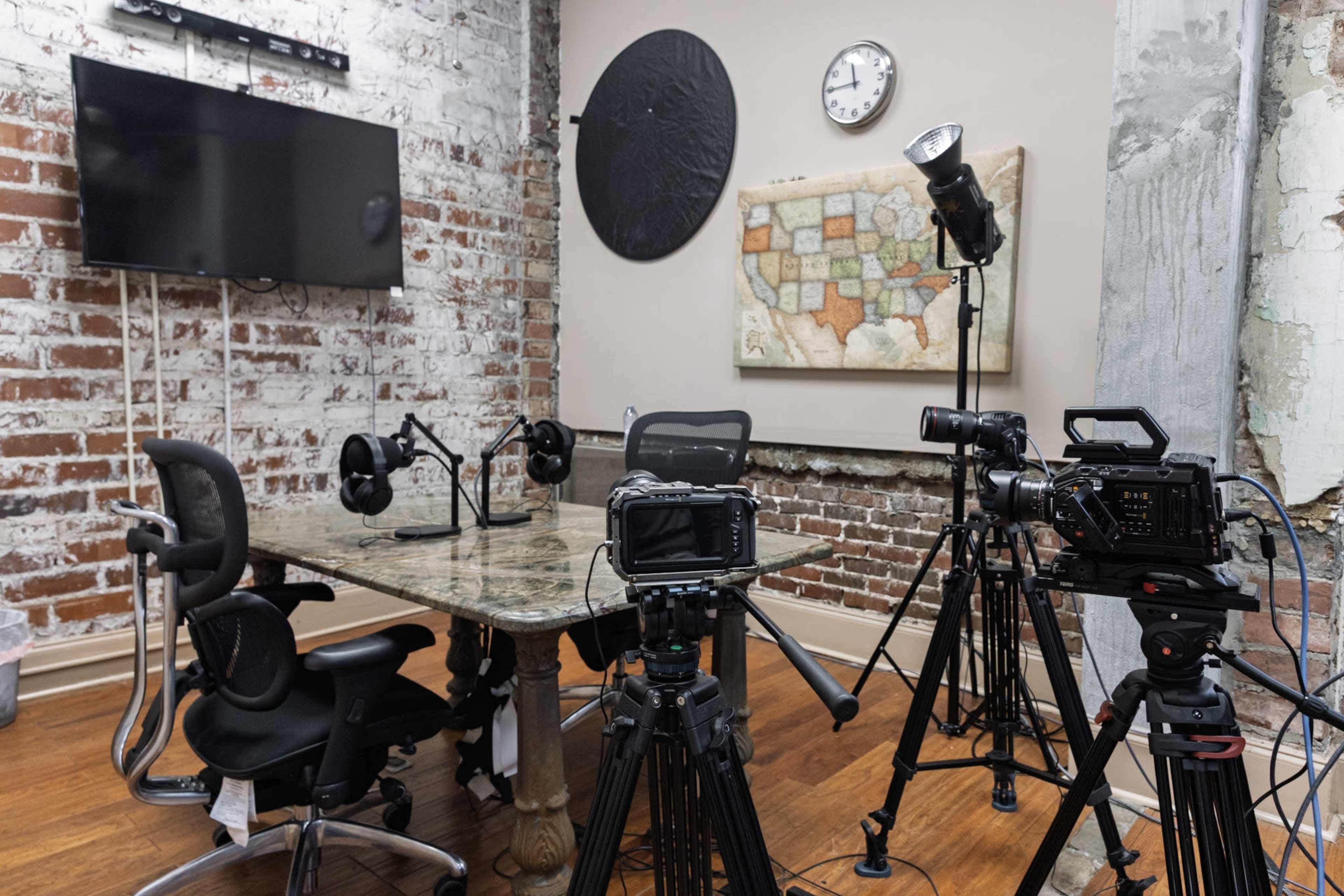 The image shows a recording studio with a table, chairs, cameras, and a large wall-mounted display in a room featuring exposed brick walls.