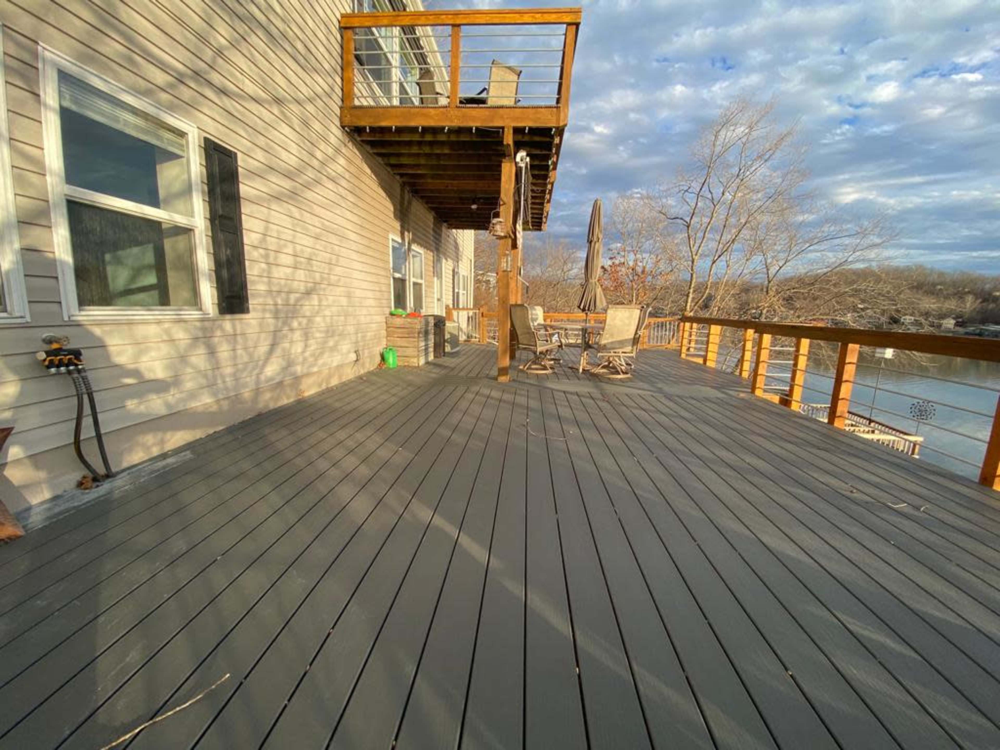 A wooden deck with a railing, featuring two chairs and a table, adjacent to a house and overlooking a body of water.