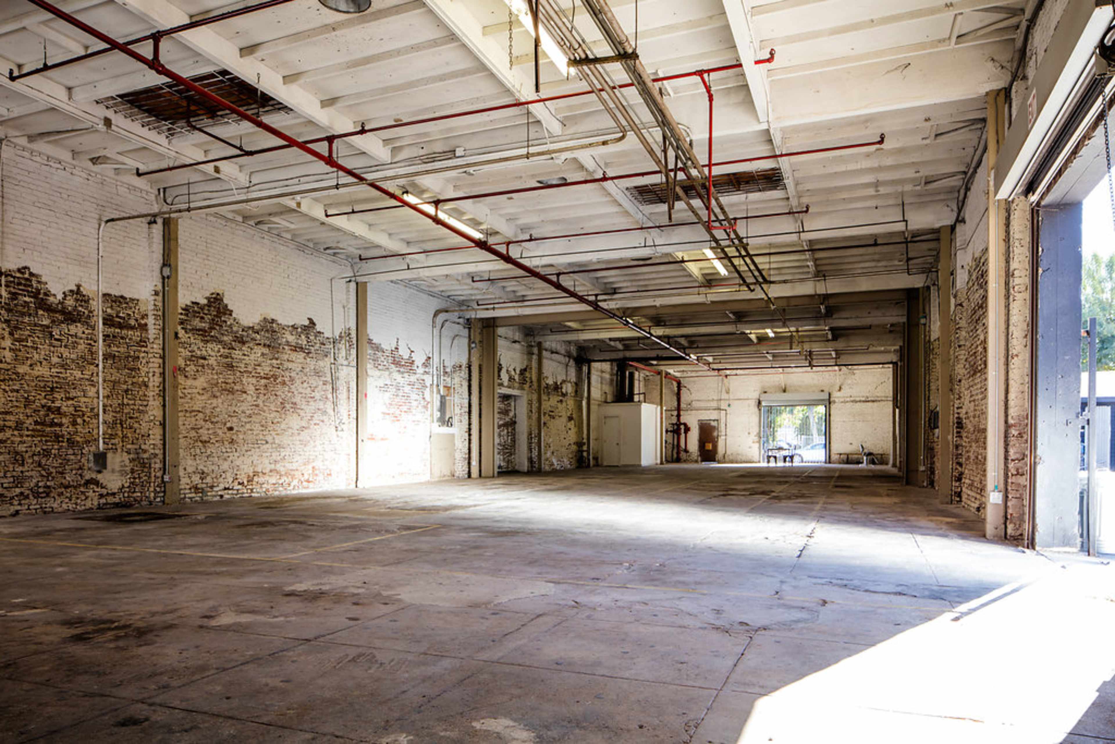 An empty industrial space features exposed brick walls, a high ceiling with pipes, and ample natural light streaming through large openings.