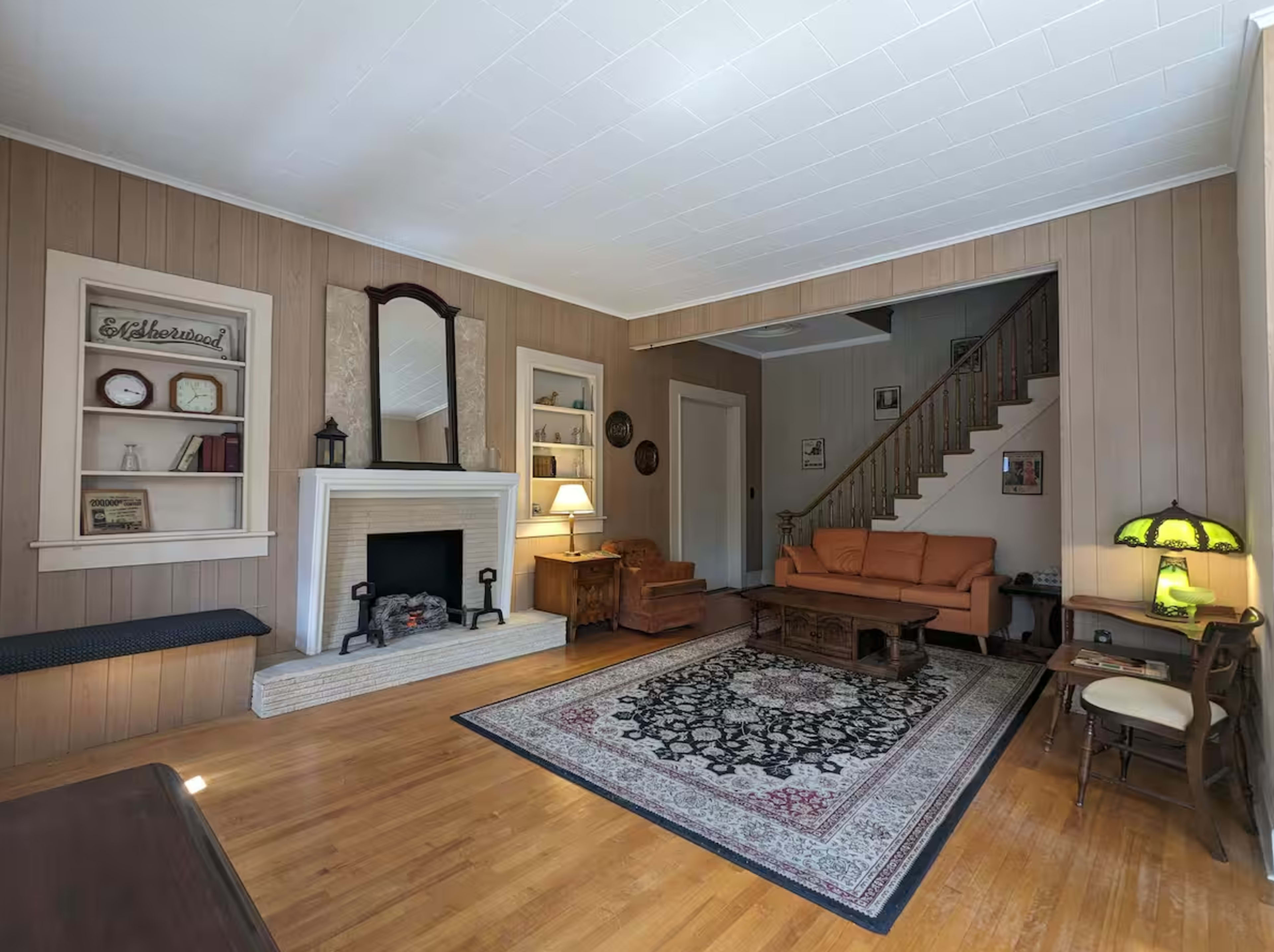 The living room features a fireplace, a staircase, and a combination of seating arrangements with a rug on hardwood flooring.