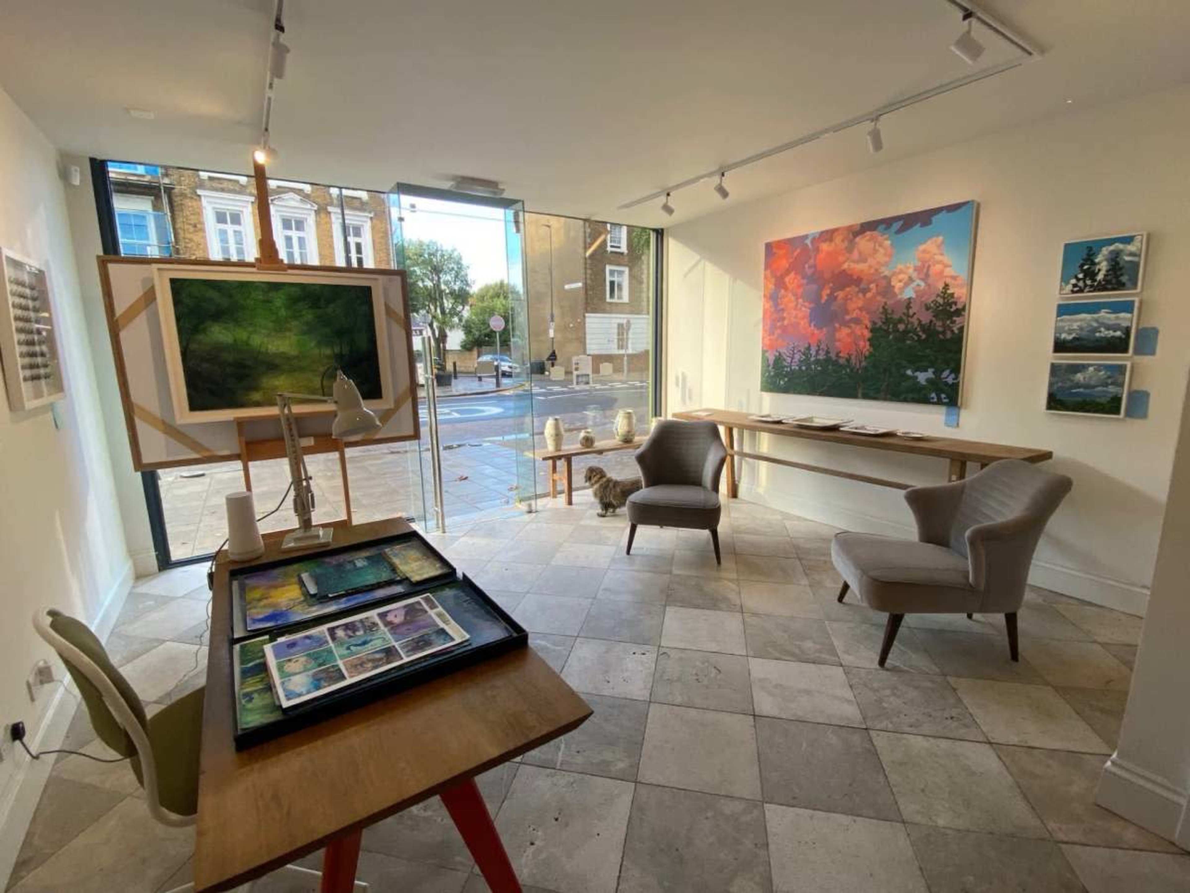 The image shows an art gallery featuring various artworks displayed on the walls and a table with art supplies in the foreground.