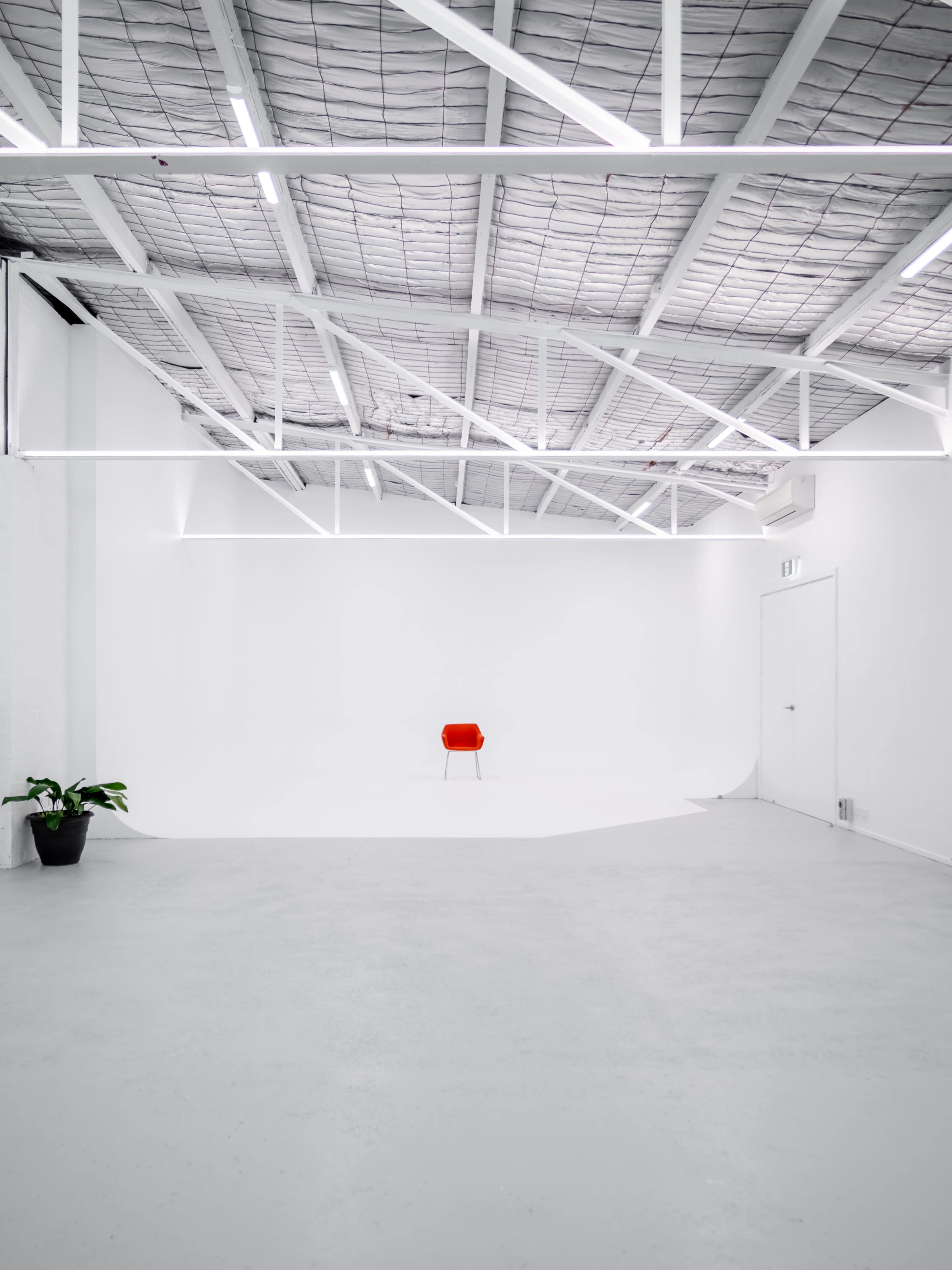 A minimalist, empty studio space with white walls, a concrete floor, and a single red chair placed in the center.