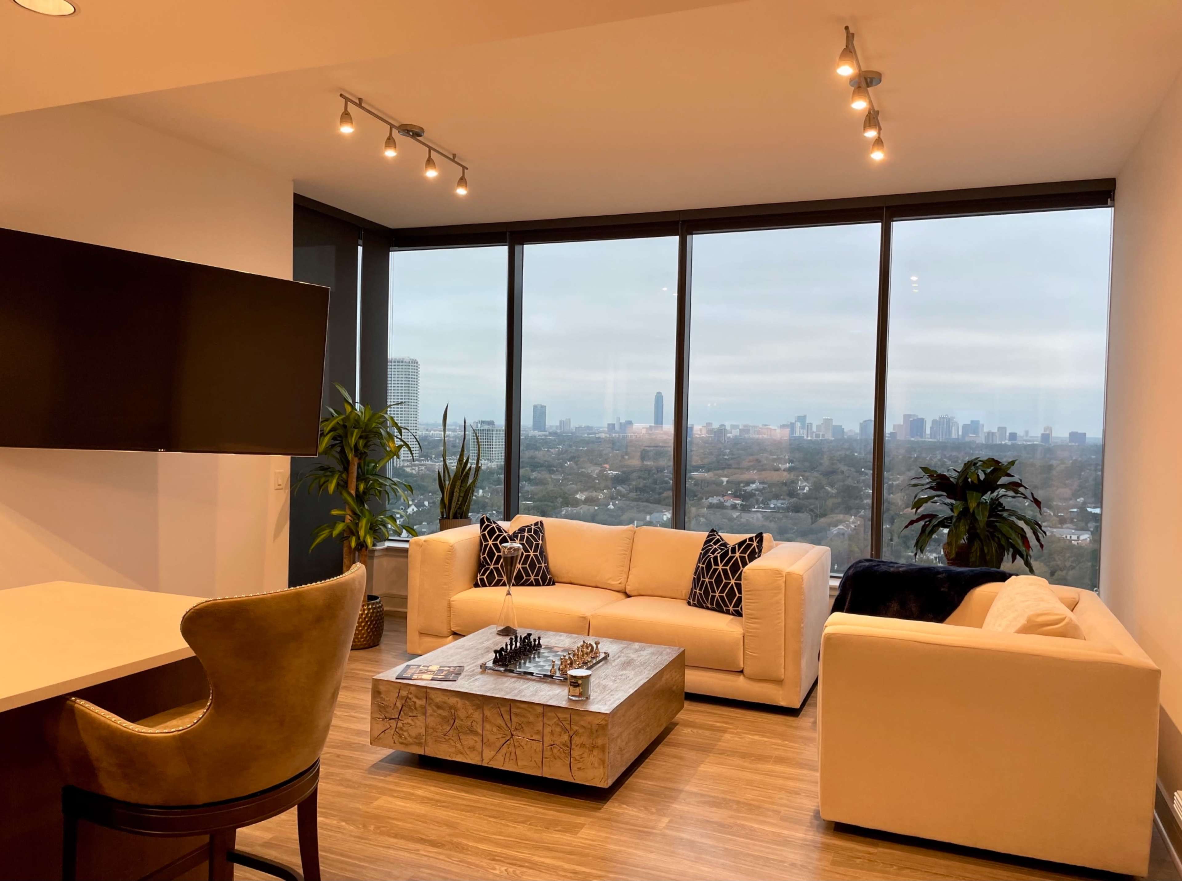 A modern living room features two sofas and a coffee table with a chess set, overlooking a city skyline through large windows.