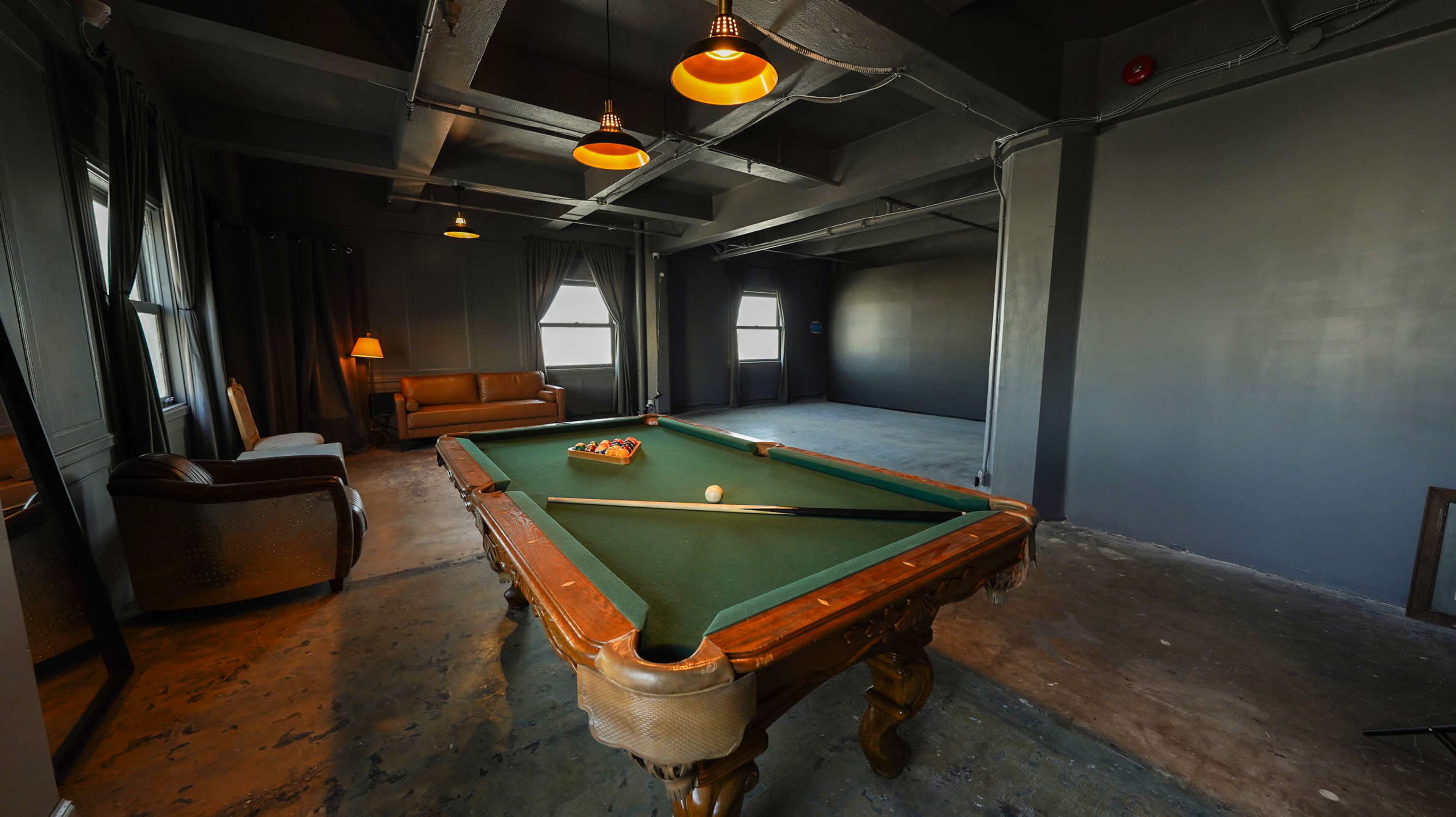 A room with a billiards table set in the center, flanked by a sofa and an armchair, with windows and ceiling lights illuminating the space.
