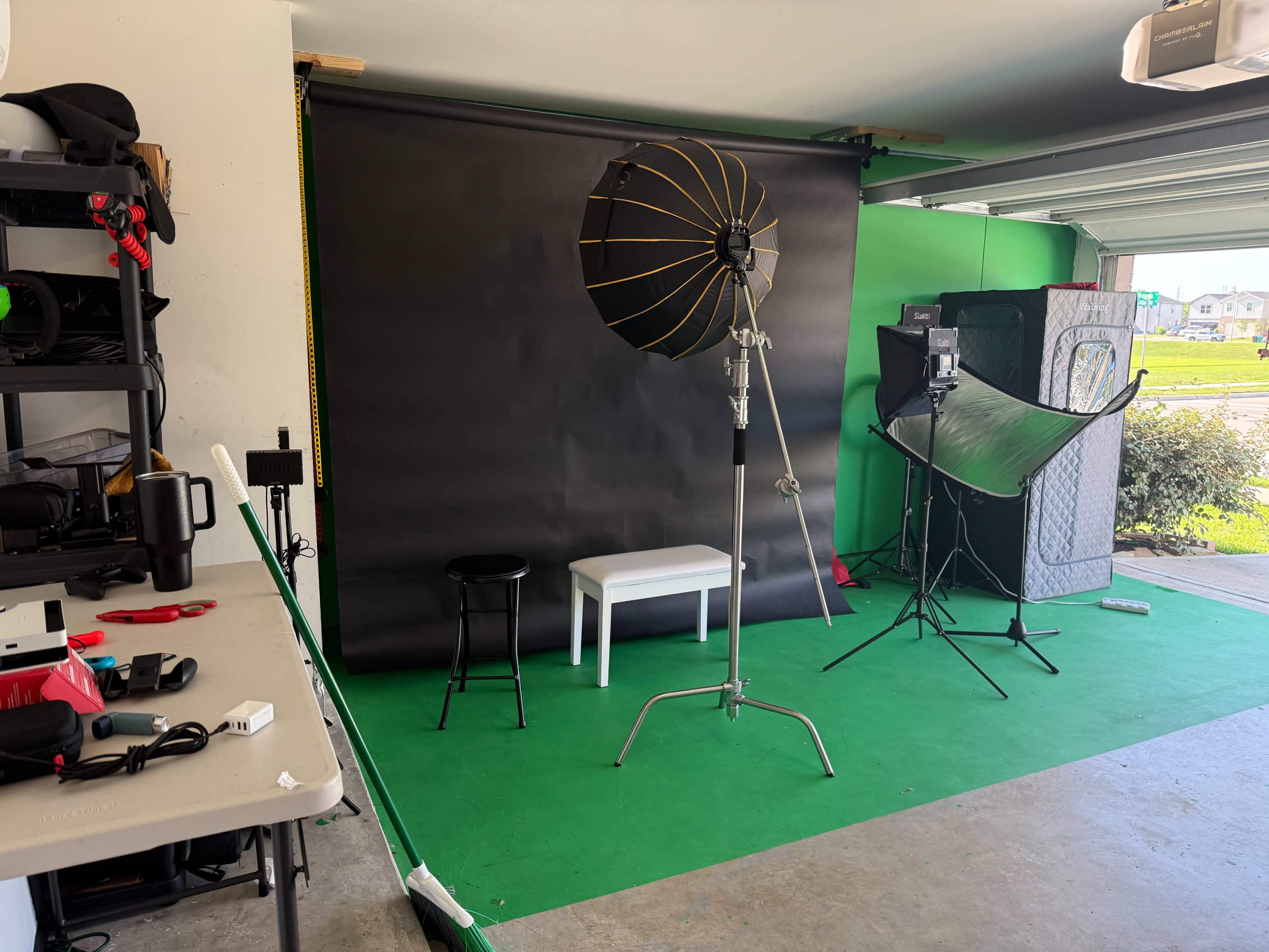 The image shows a photography studio setup with a green backdrop, a white stool, a black stool, and various lighting equipment arranged in a garage.