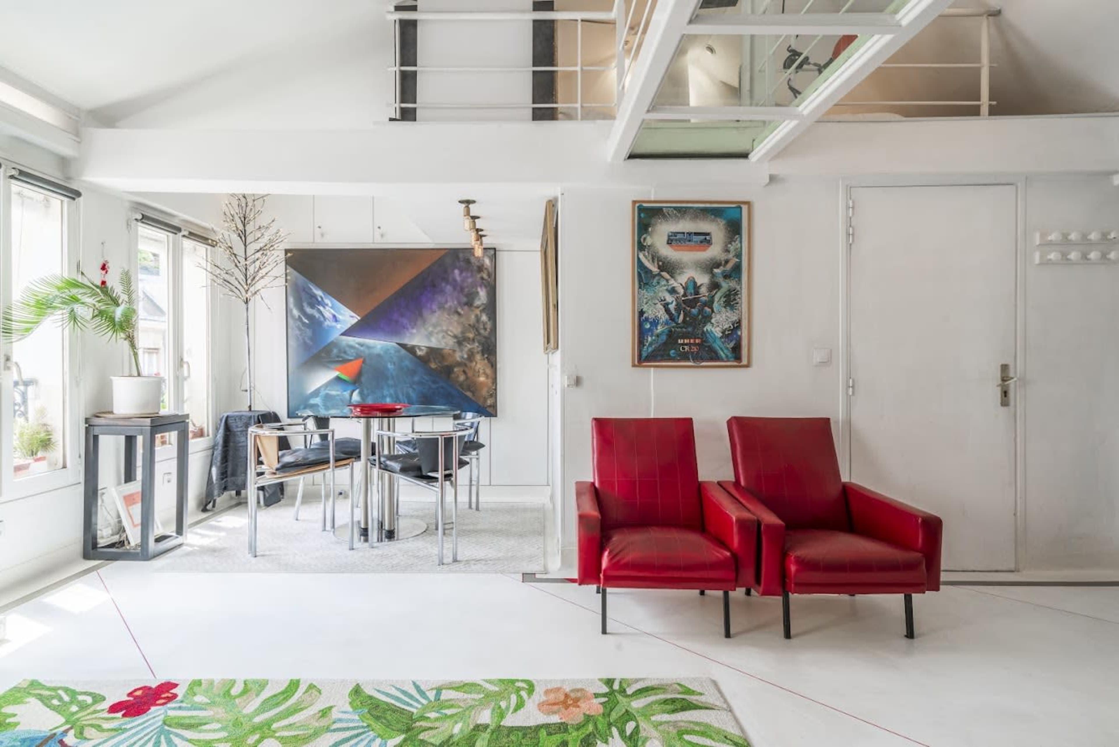 The image shows a bright interior space featuring two red armchairs, a glass dining table with chairs, and a large abstract painting on the wall.