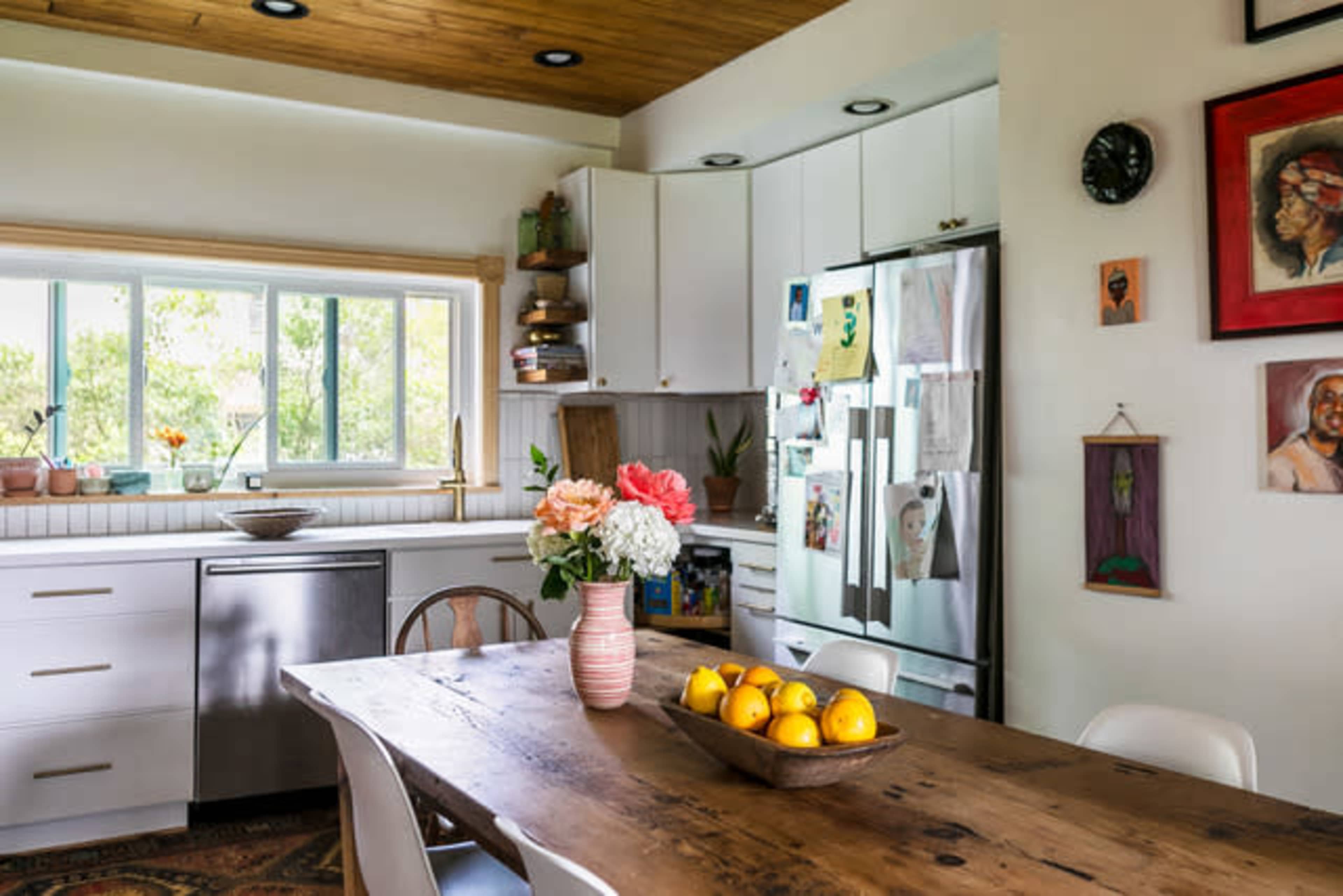 A bright kitchen features a wooden dining table with a bowl of lemons, surrounded by white chairs and cabinets, with art on the walls and a large window letting in natural light.